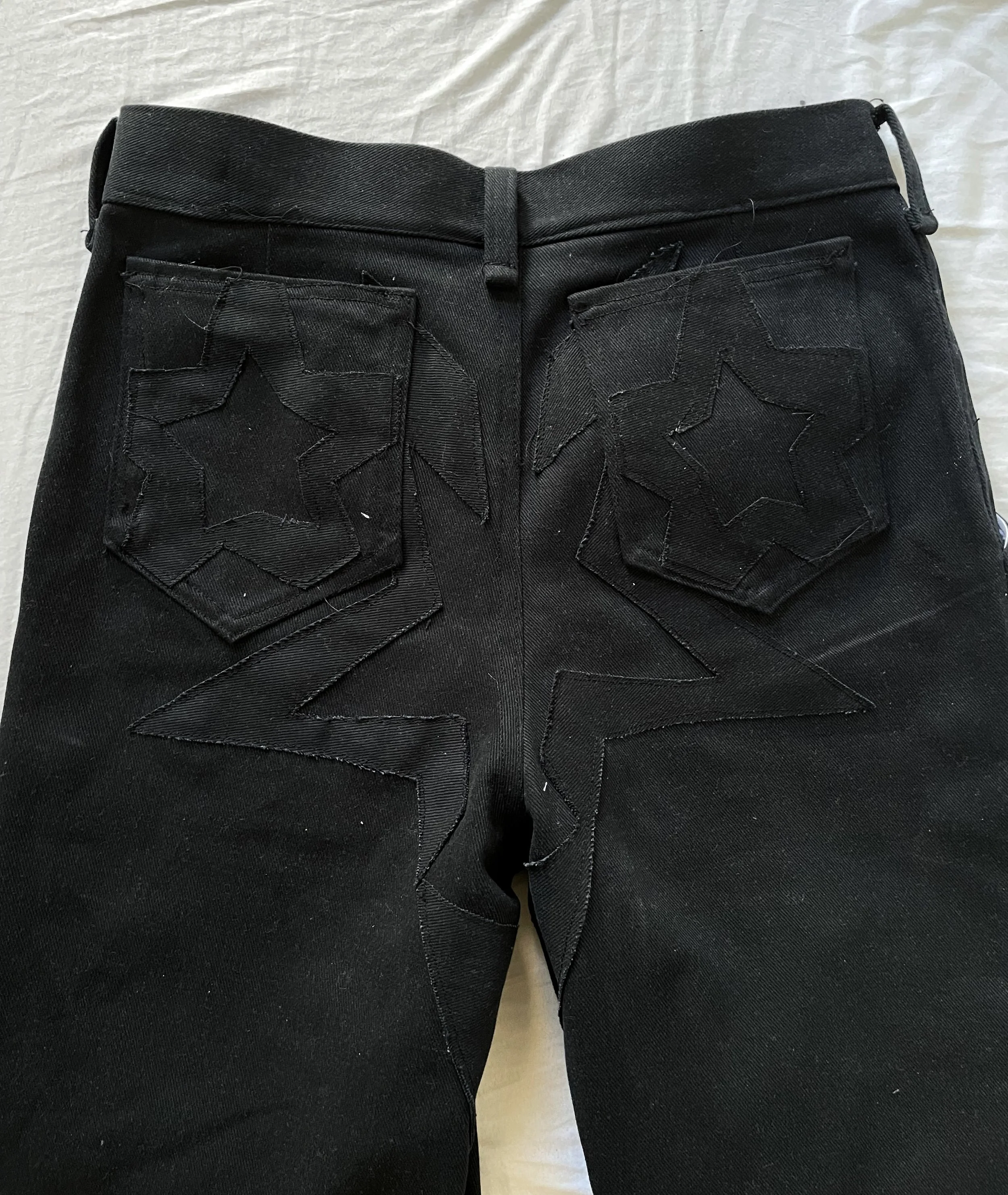 Black jeans with star-shaped stitched patches on the back pockets, lying on a white surface. conor o'driscoll
