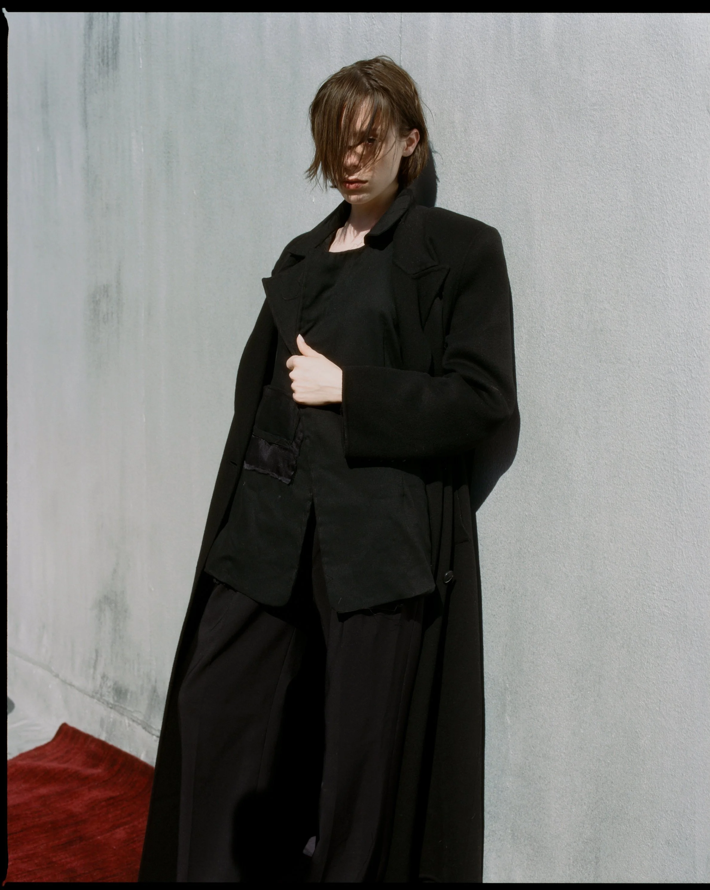 Ella Snyder standing against a light gray wall, dressed in an all-black outfit including a long coat, shirt, and wide-legged pants. They have shoulder-length brown hair partially covering their face and are looking downward with a serious expression.