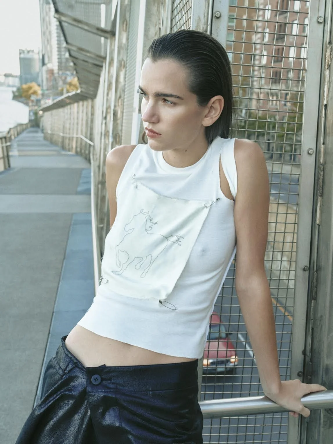 A woman with dark, slicked-back hair leaning against a metal railing on a bridge or walkway, with a cityscape and water in the background, wearing a white sleeveless crop top with a stitched unicorn outline on it and black shiny pants.