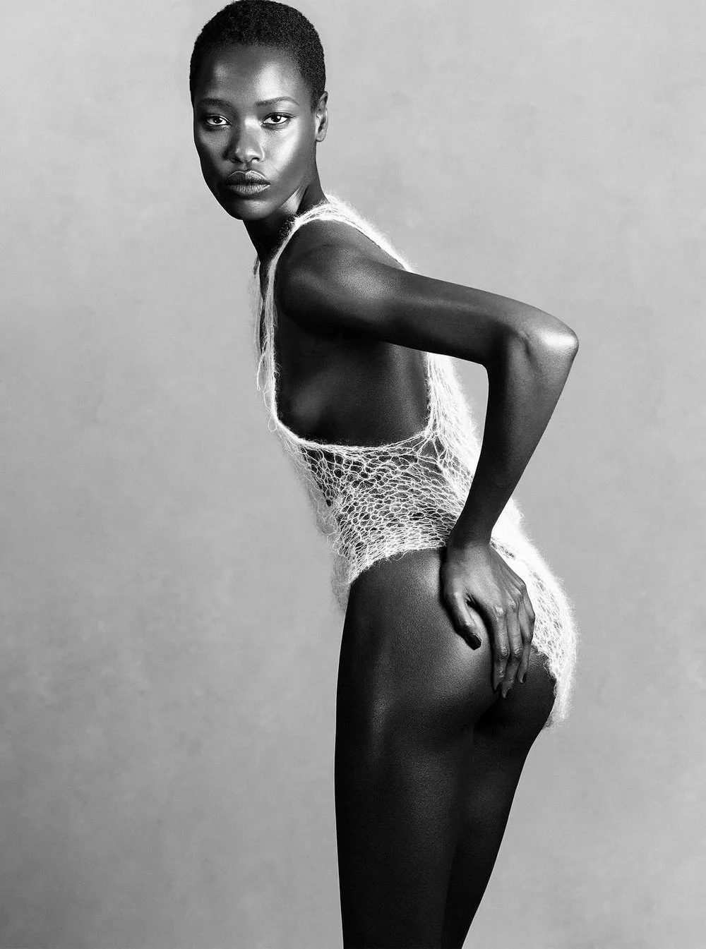 Black and white photograph of a young woman with a short hairstyle, wearing a delicate, loose-knit top, posing with one hand on her hip and looking directly at the camera. conor o'driscoll