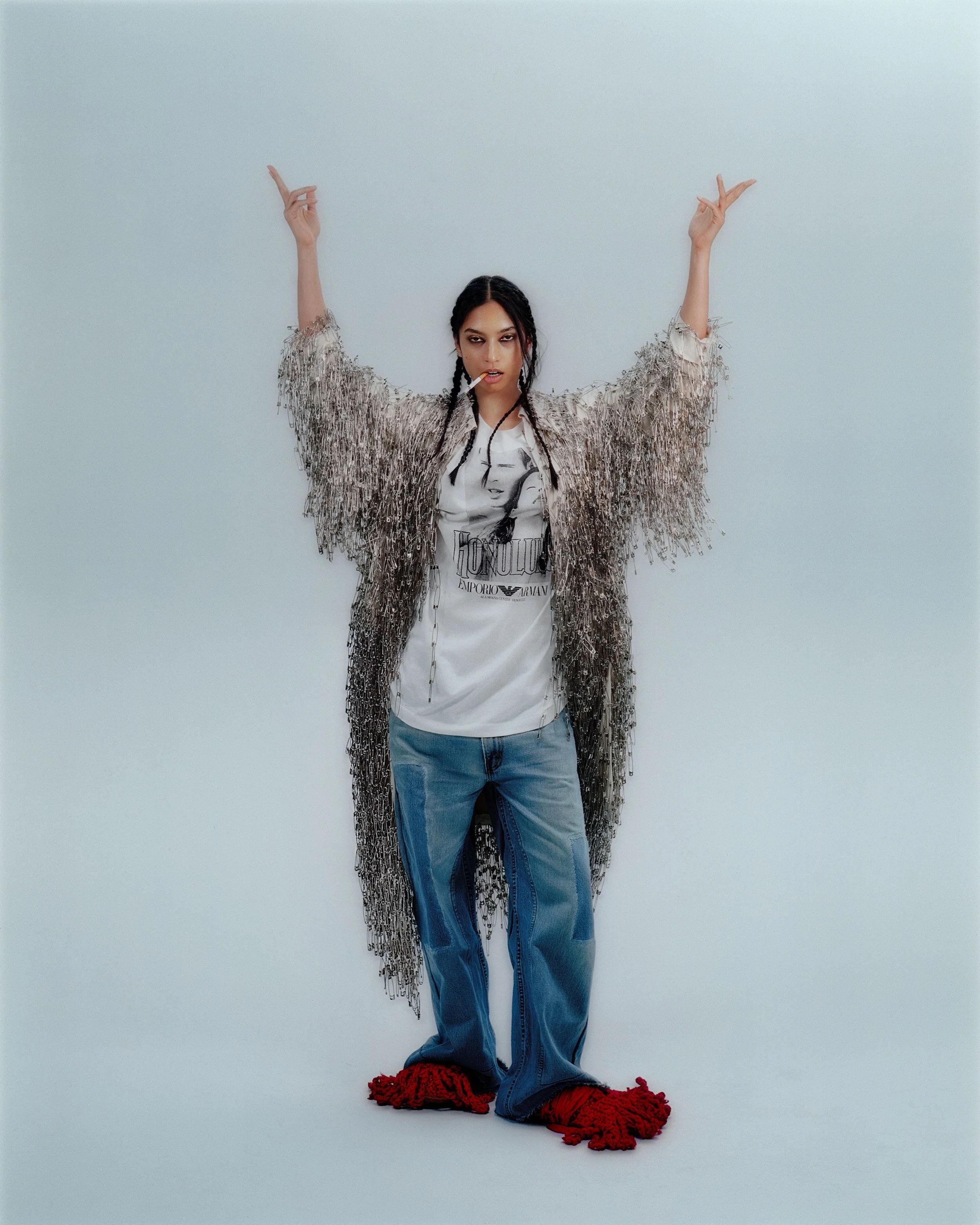 A woman with dark hair and bold makeup posing with arms raised and hands pointing upward, dressed in a graphic t-shirt, jeans, a large fringed jacket, and red slippers, standing against a plain wall.CONOR O'DRISCOLL