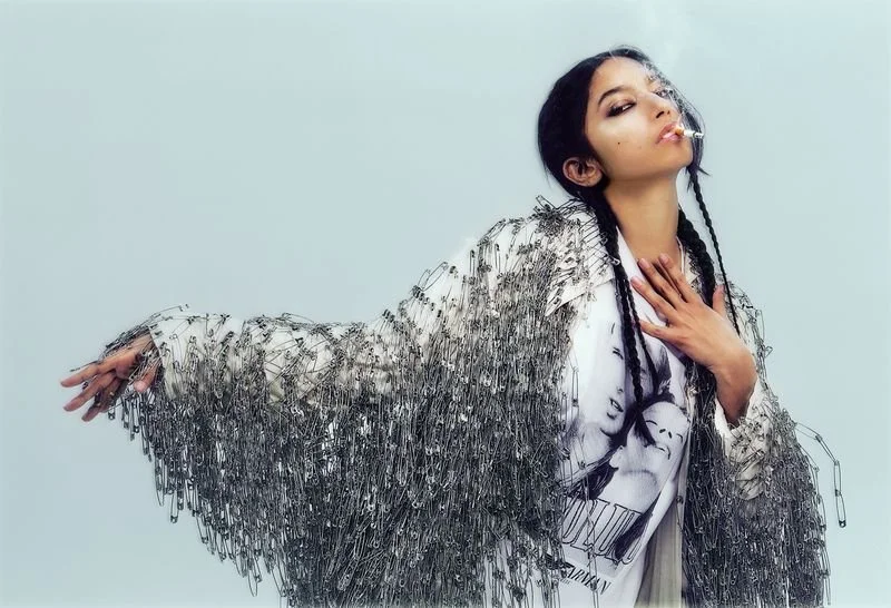 A woman with long, dark hair styled in braids, wearing a safety pin coat, a graphic t-shirt, and holding a cigarette in her mouth. CONOR O'DRISCOLL