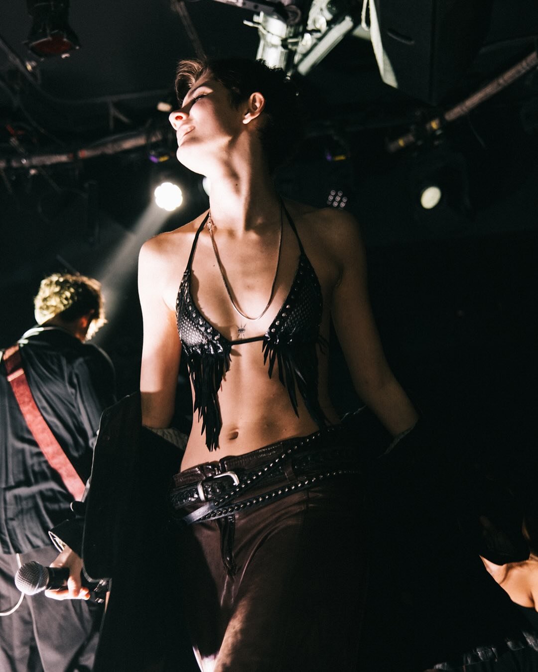 A woman with short dark hair wearing a black fringed top, black pants with a chain belt, and multiple necklaces, holding a microphone, performing on stage with a man in the background and stage lighting overhead. by conor o'driscoll