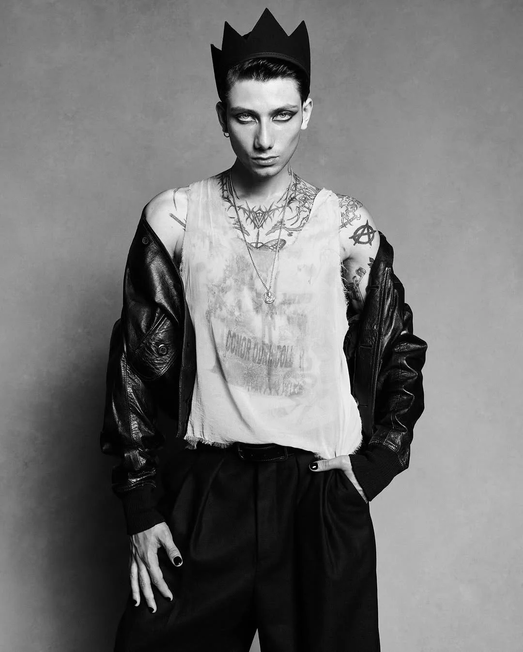 A person with short dark hair, wearing a crown-shaped headband, a sleeveless graphic t-shirt, a leather jacket draped over shoulders, and black pants. They have tattoos on their chest, arms, and neck, and are posing with a confident Conor O'Driscoll