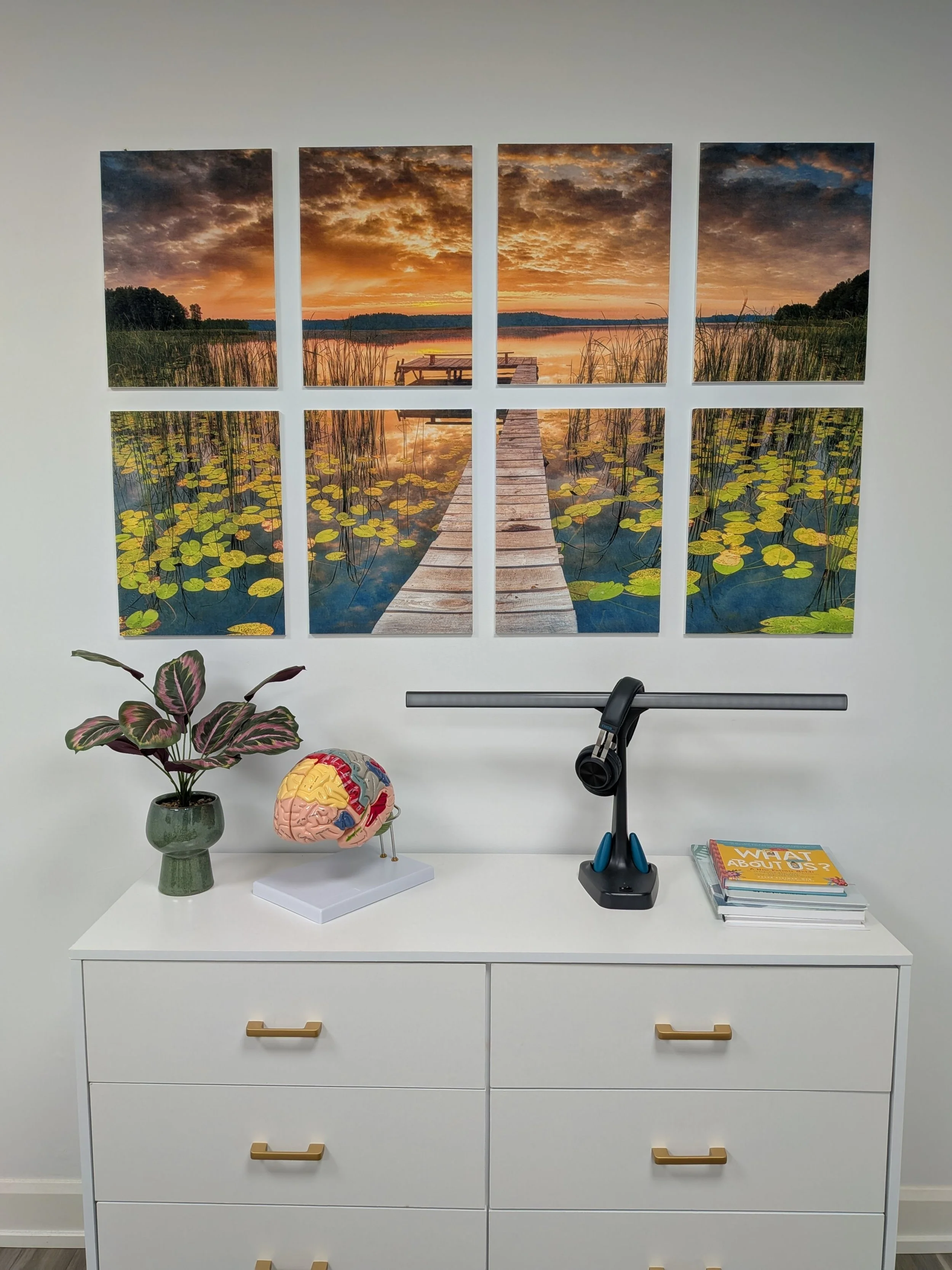 White dresser with gold handles, decorated with a potted plant, a model of a human brain, a gimbal, and a stack of colorful books. Above, a multi-panel wall art of a lake scene at sunset with a wooden dock extending into the water surrounded by lily 