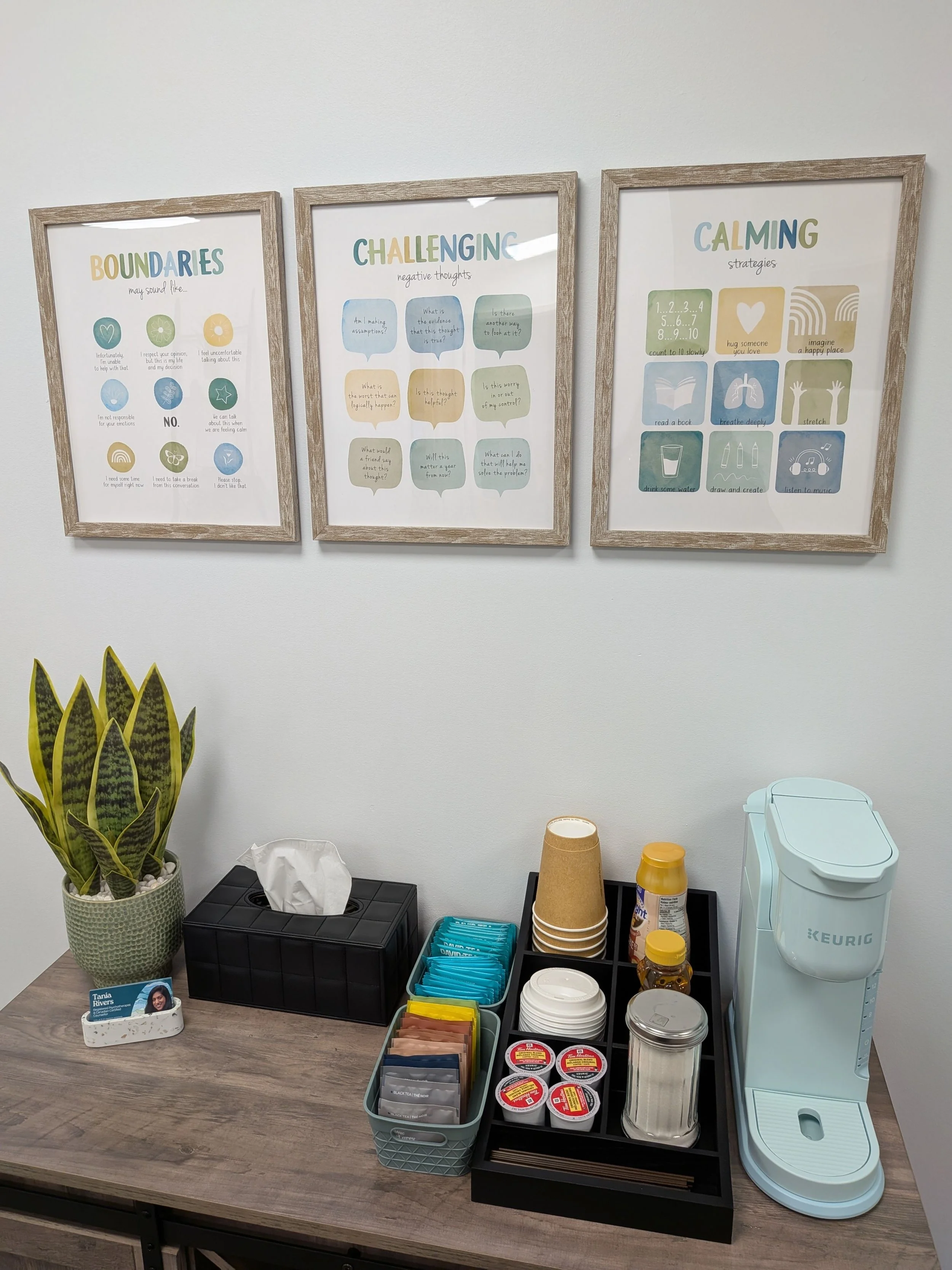 Document on a countertop with a pot of snake plant, a black tissue box, a container of sugar and creamer pods, disposable cups, and a Keurig coffee machine. Three framed posters titled 'Boundaries', 'Challenging negative thoughts', and 'Calming strat