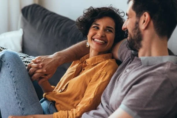 A woman and man sitting on a couch, smiling and holding hands, engaged in a happy moment.