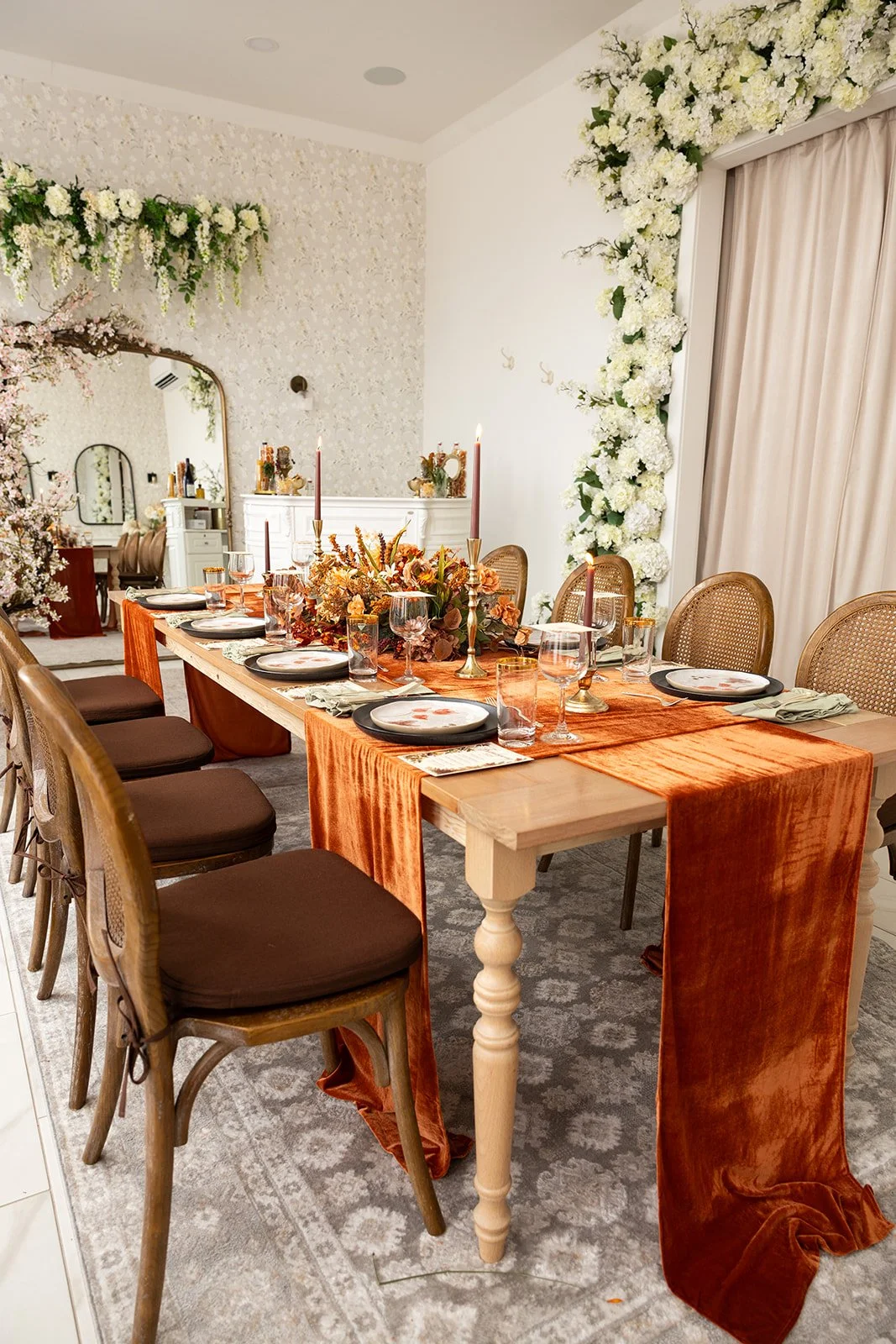 An elegant styled Friendsgiving tablescape at The Lovely on Main, featuring rich burnt orange velvet table runners, lush autumn floral centerpieces, glowing taper candles, botanical dinnerware, sage green napkins, and vintage-inspired glassware.