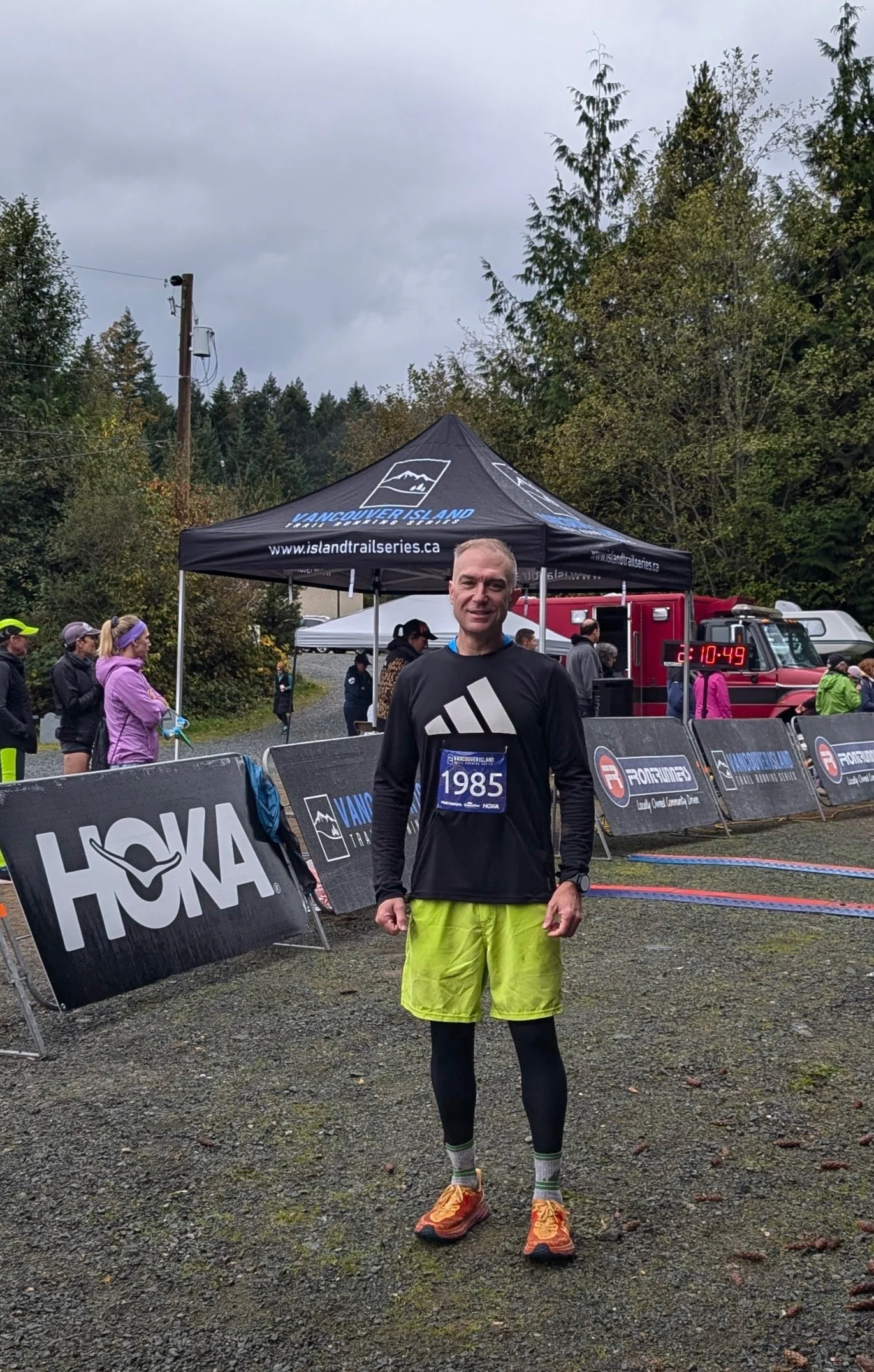 Male runner at the finish line of a trail race, wearing a black long-sleeve shirt, bright yellow shorts, black leggings, and orange running shoes, standing under a canopy with other runners and a digital timer in the background.