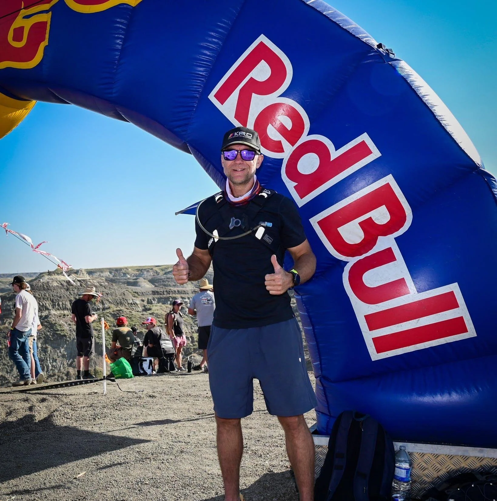 Man standing under a Red Bull arch at an outdoor event, with people in the background, some wearing hats, and a dry, canyon landscape under a clear blue sky.