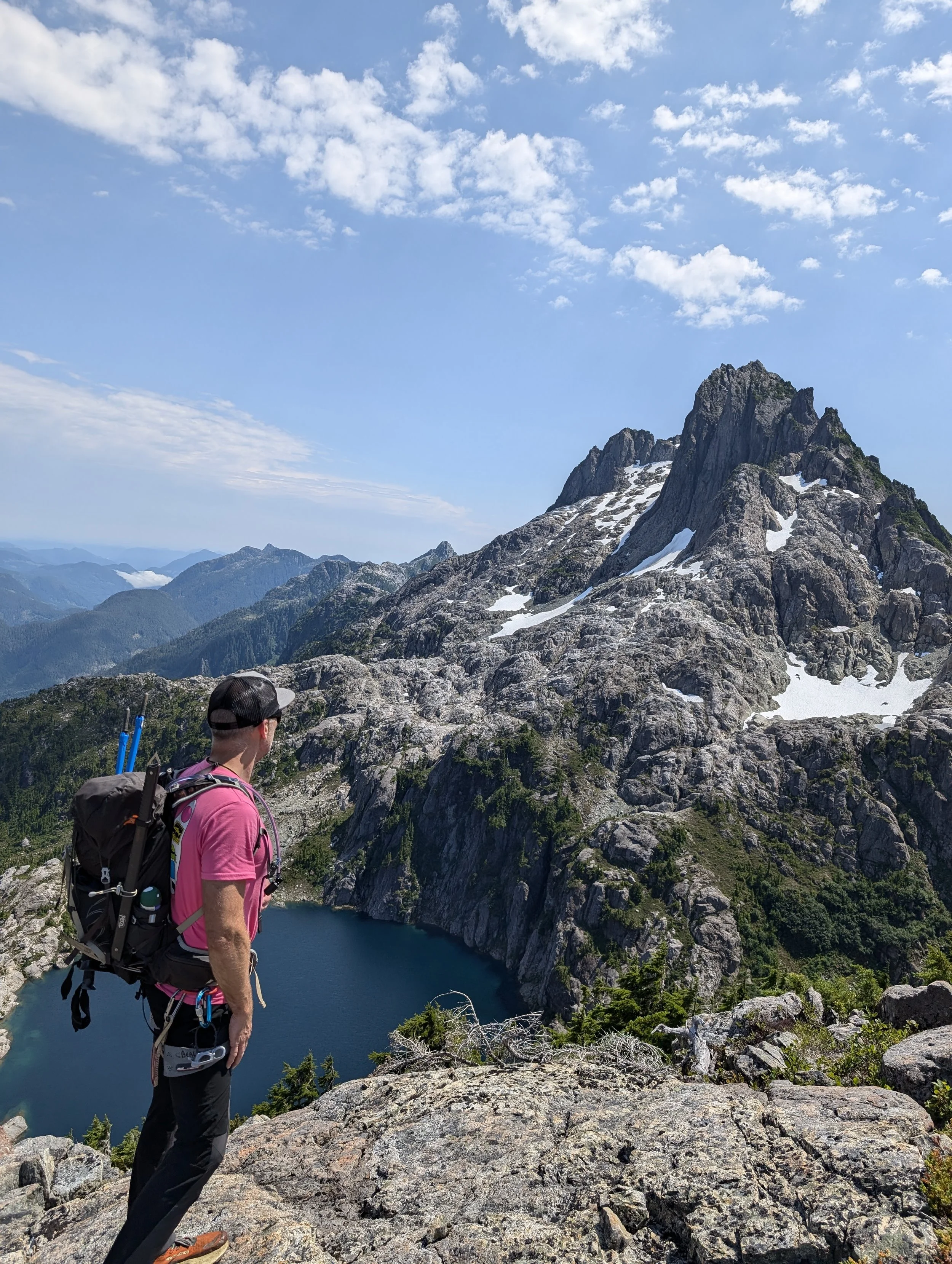 Hiker in a pink shirt with a backpack and trekking poles overlooking a mountain lake under a partly cloudy sky.