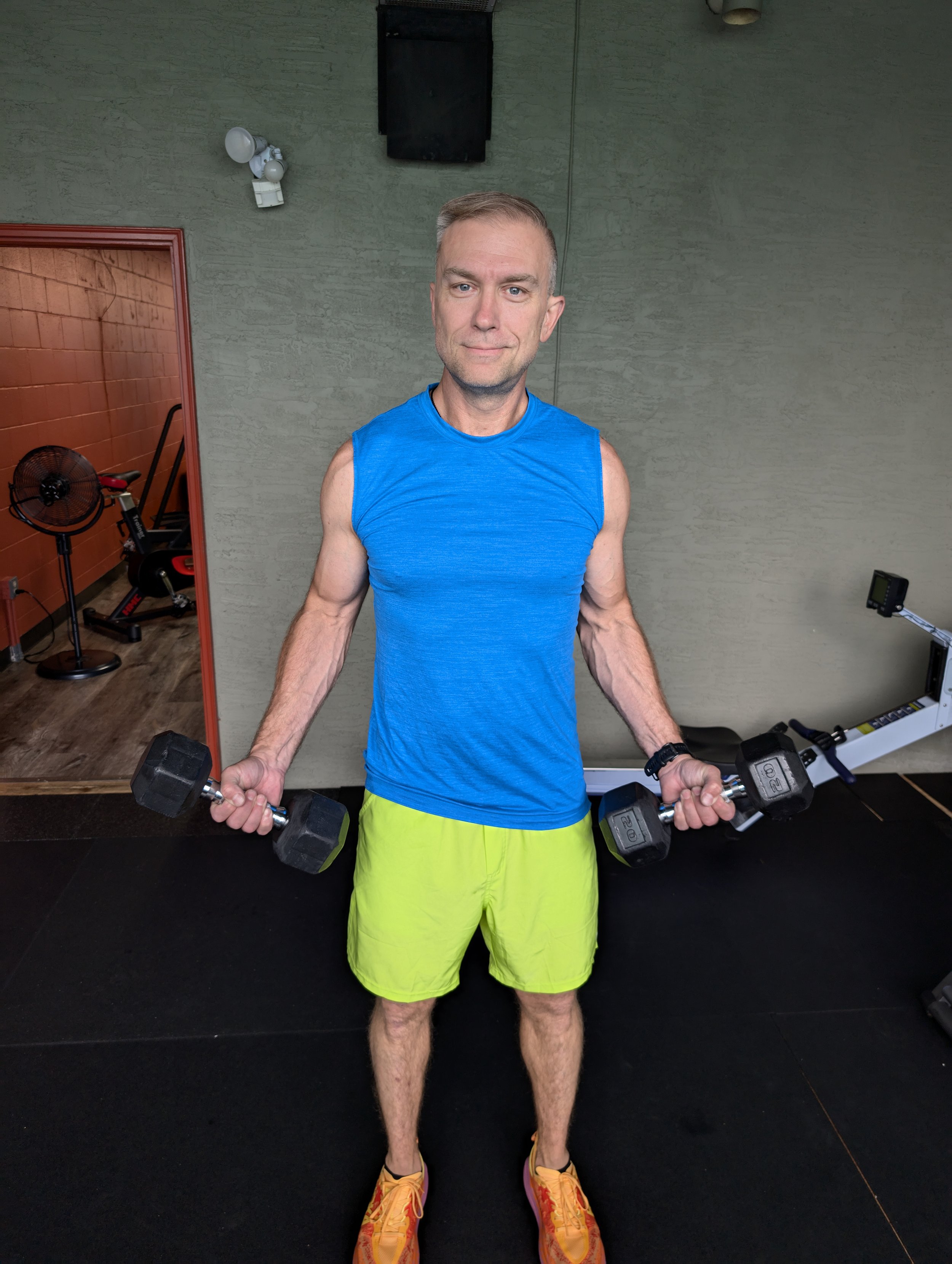 A man working out with dumbbells in a gym, wearing a blue sleeveless shirt, neon yellow shorts, and orange athletic shoes.