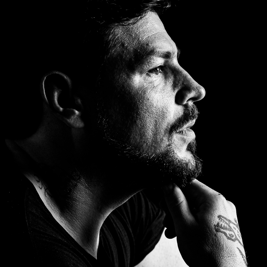Black and white close-up side profile of a man with a beard, looking contemplative, with his hand resting on his chin and a tattoo on his wrist.