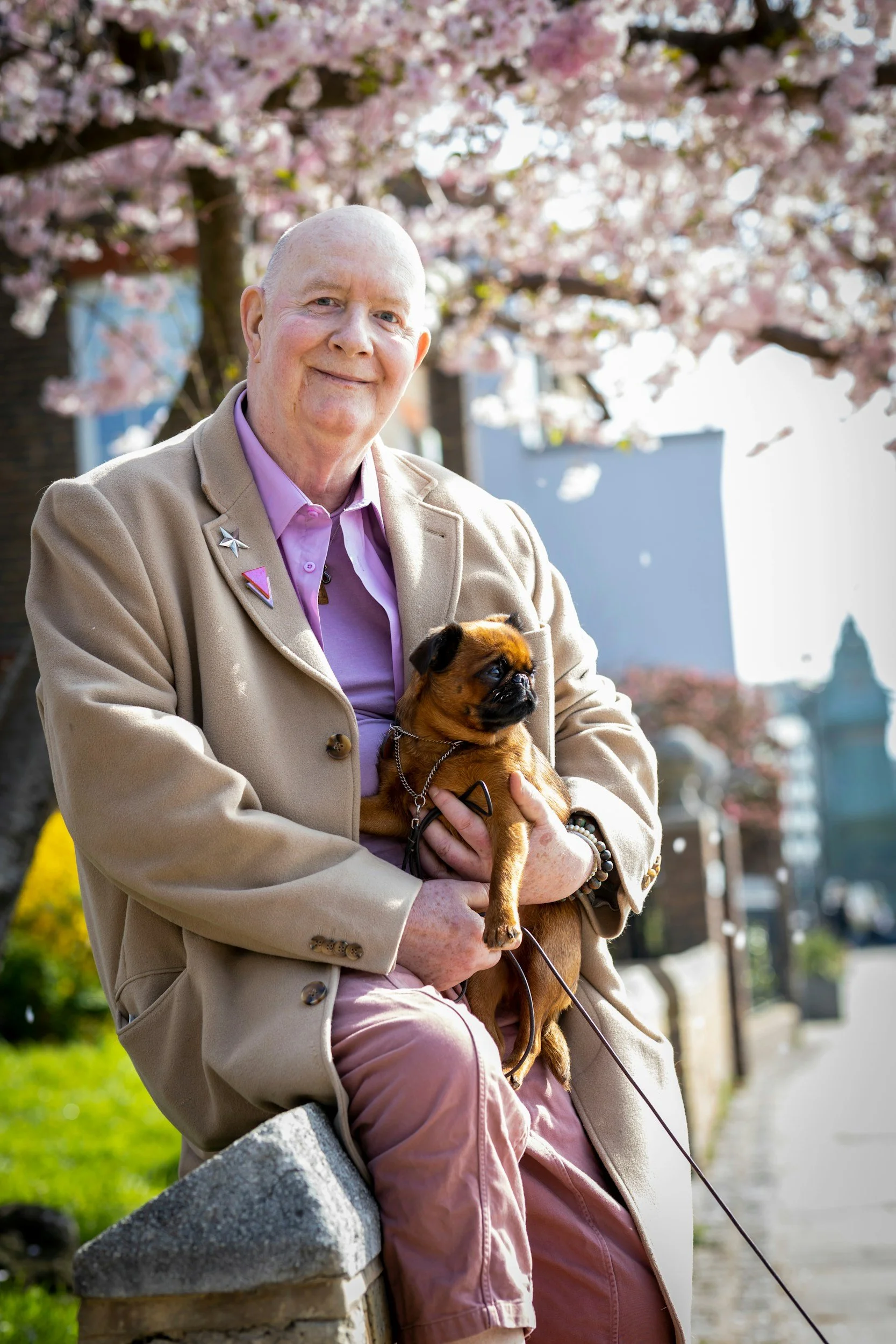 An elderly man smiling and sitting on a stone bench outdoors, holding a small brown dog in his lap. The man wears a beige coat and pink shirt with several pins on the lapel, and the background shows a flowering tree with pink blossoms.