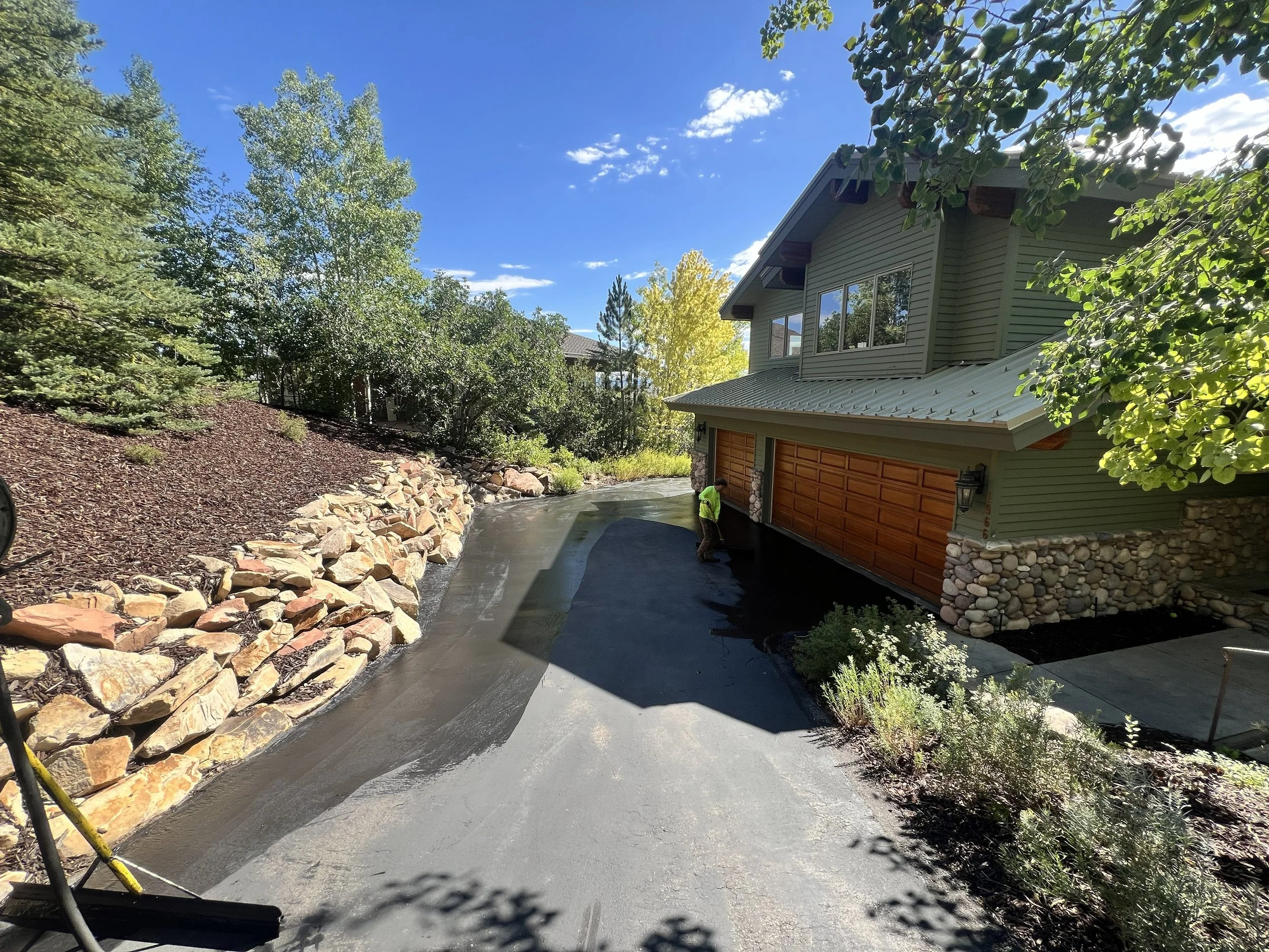 Worker applying driveway sealcoat on a residential asphalt driveway  surface beside a landscaped rock wall area
