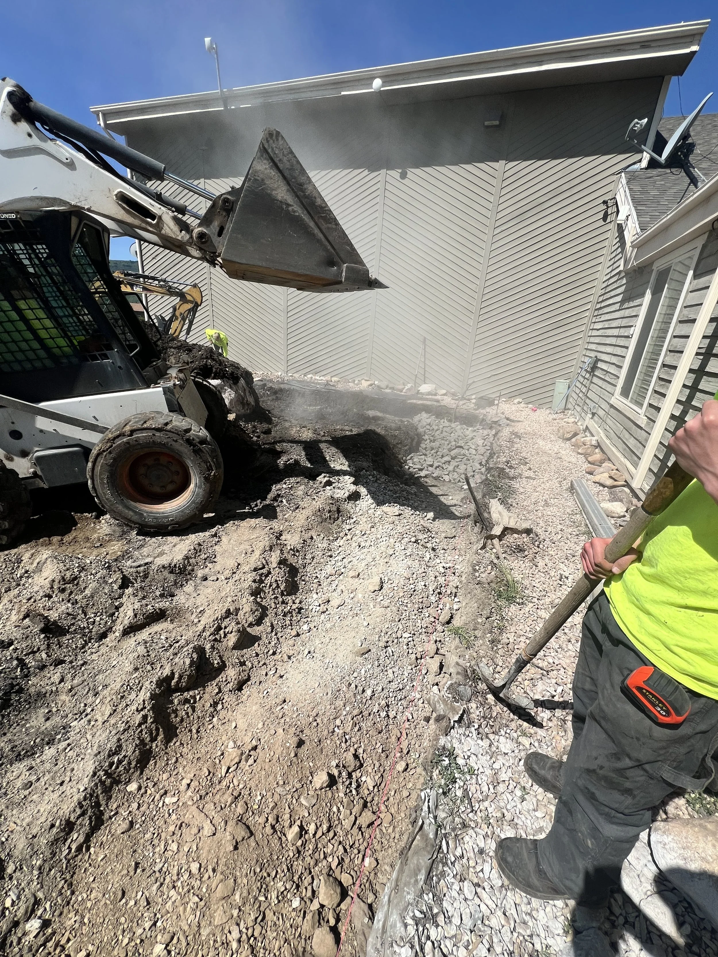 Construction worker with a shovel working on a dirt and gravel area near a house, with a backhoe loader excavator in the background.