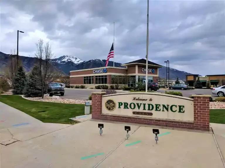 Welcome to Providence City sign in Providence Utah with Chase Bank in the background, an overcast sky, and snow‑capped mountains in the far distance, near residential and commercial areas served by asphalt paving, repair, and maintenance services.