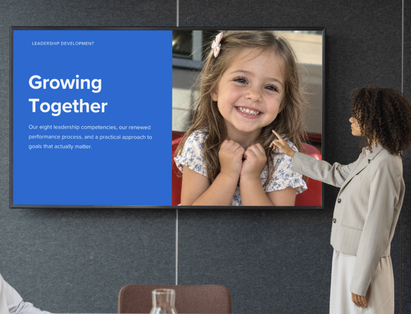 A woman in a gray blazer points at a large screen displaying a smiling young girl with long brown hair. The screen shows a blue section with white text about leadership development and a photo of the girl with her hands clasped, smiling.