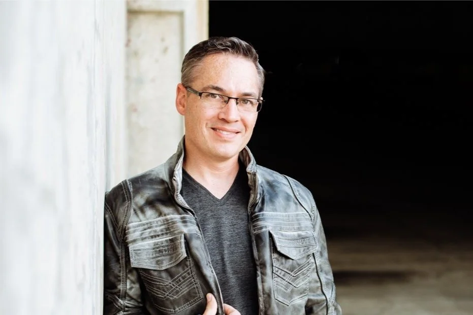 A man with glasses and short, styled hair smiling while leaning against a light-colored wall in a black leather jacket and dark shirt.