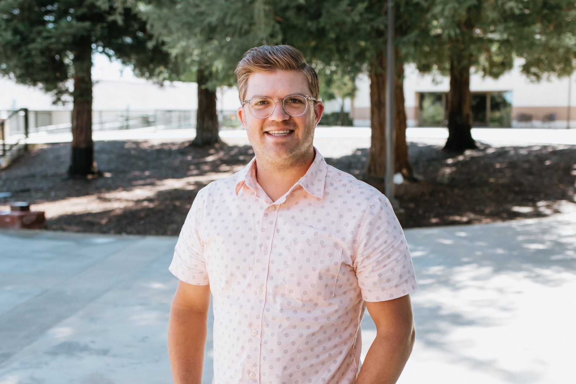Young man with glasses smiling outdoors on a sunny day, standing on a concrete path with trees in the background.