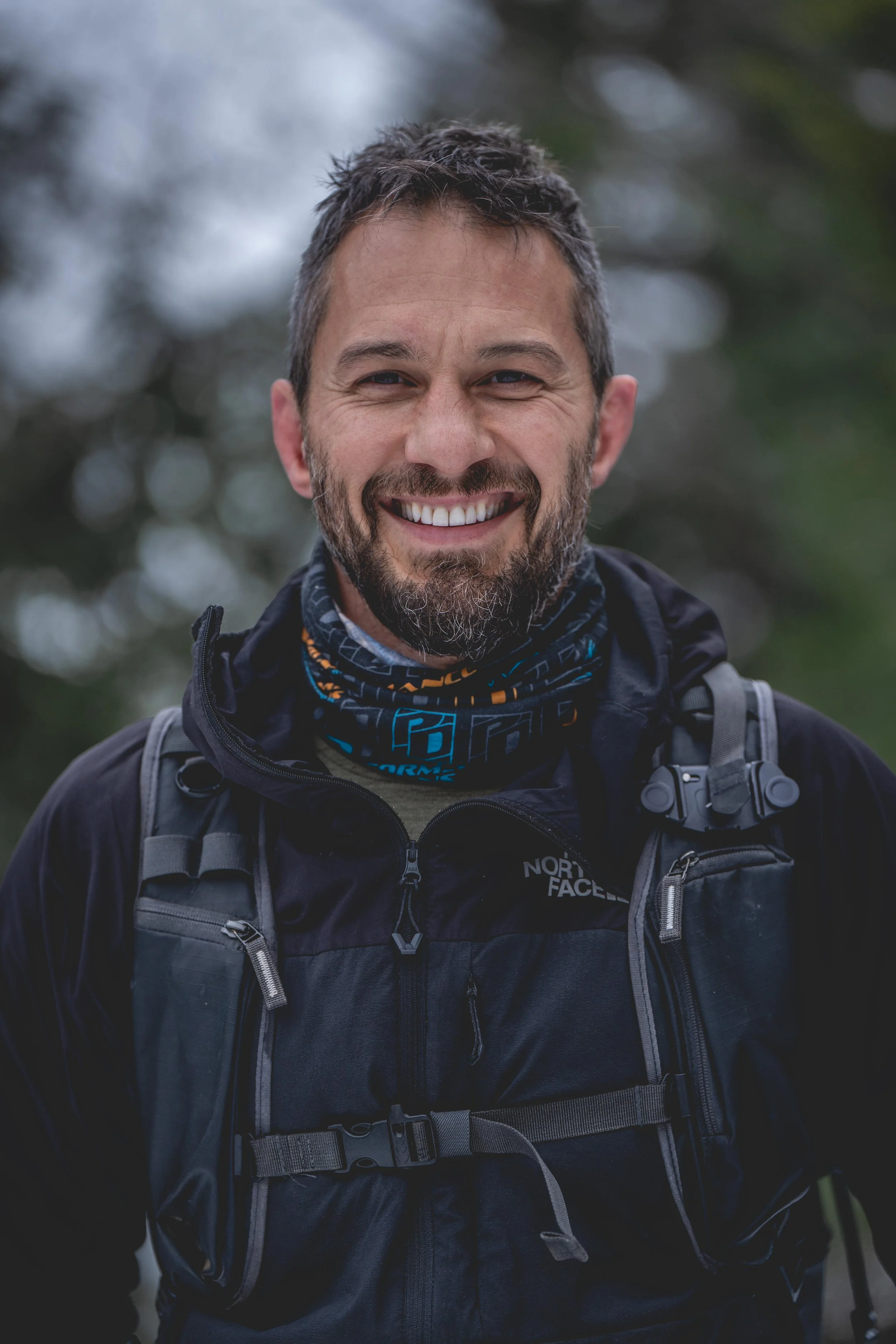 Smiling man with a beard wearing a black North Face jacket and a backpack outdoors, blurred trees in the background.