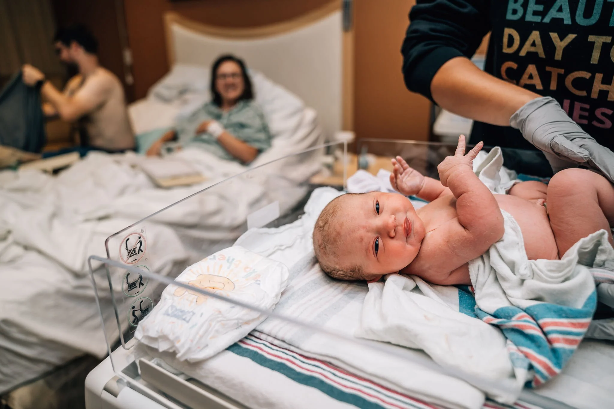 A baby lies in a warmer at a birth center while a nurse does the newborn exam with the parents watching from the bed.