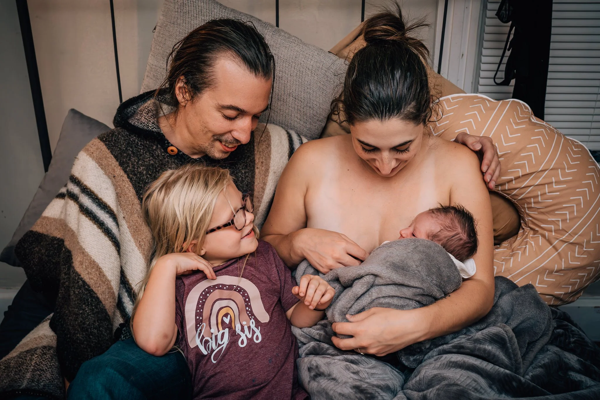 An older sibling meets their newborn sibling, while a parent is doing skin to skin in bed, after a home birth. 