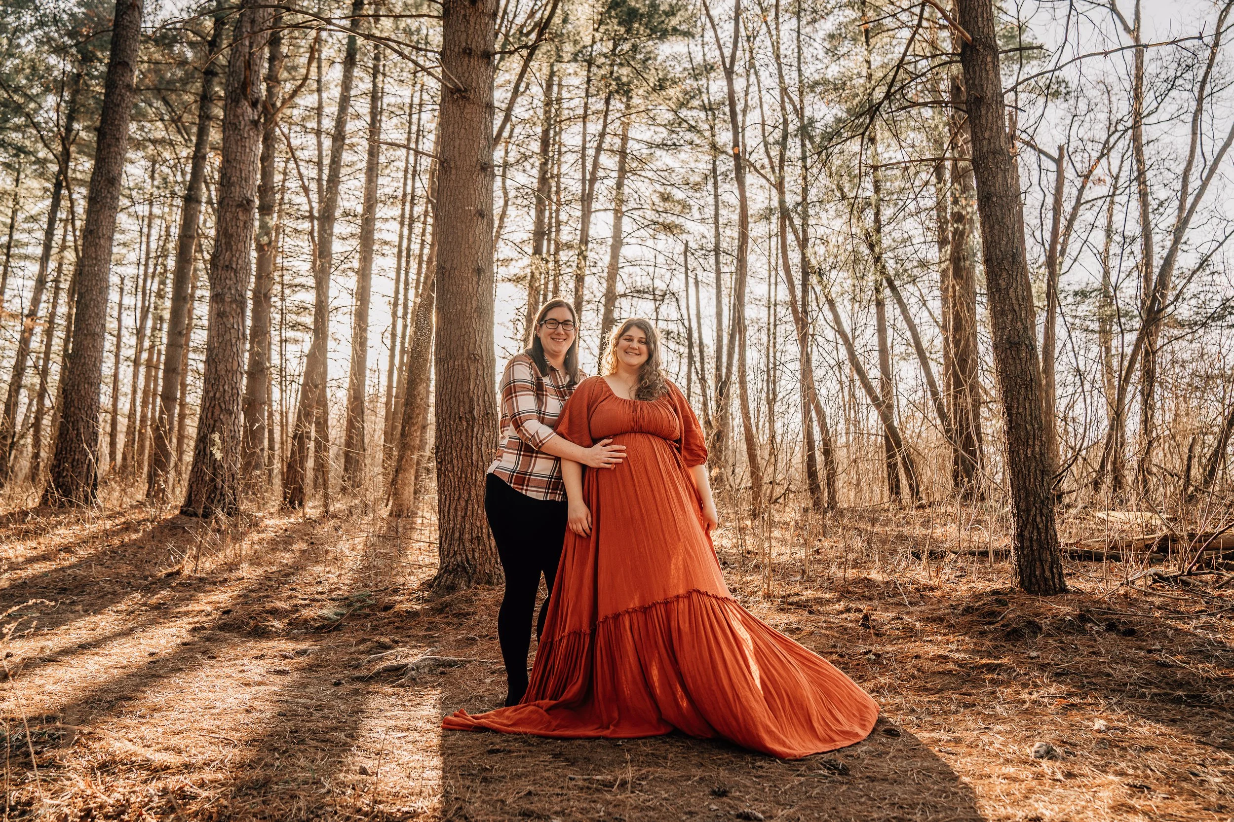 A pregnant woman in a flowing rust-colored gown stands in a sun-dappled pine forest, smiling warmly with her wife beside her. Her wife, wearing a plaid shirt and black pants, has one arm around her and the other gently resting on her belly. The soft
