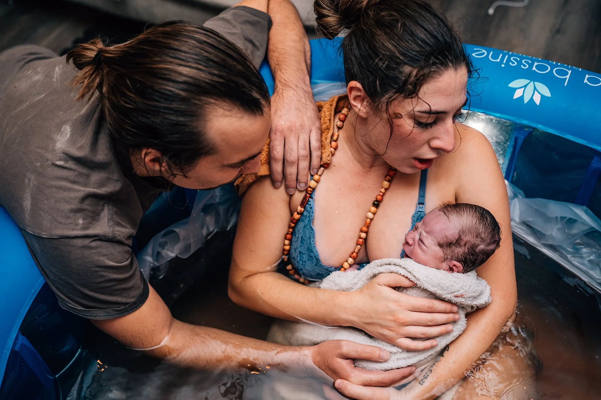 A mother holds her newborn skin to skin in a birth pool just moments after birth. The father leans in, embracing both mother and baby.