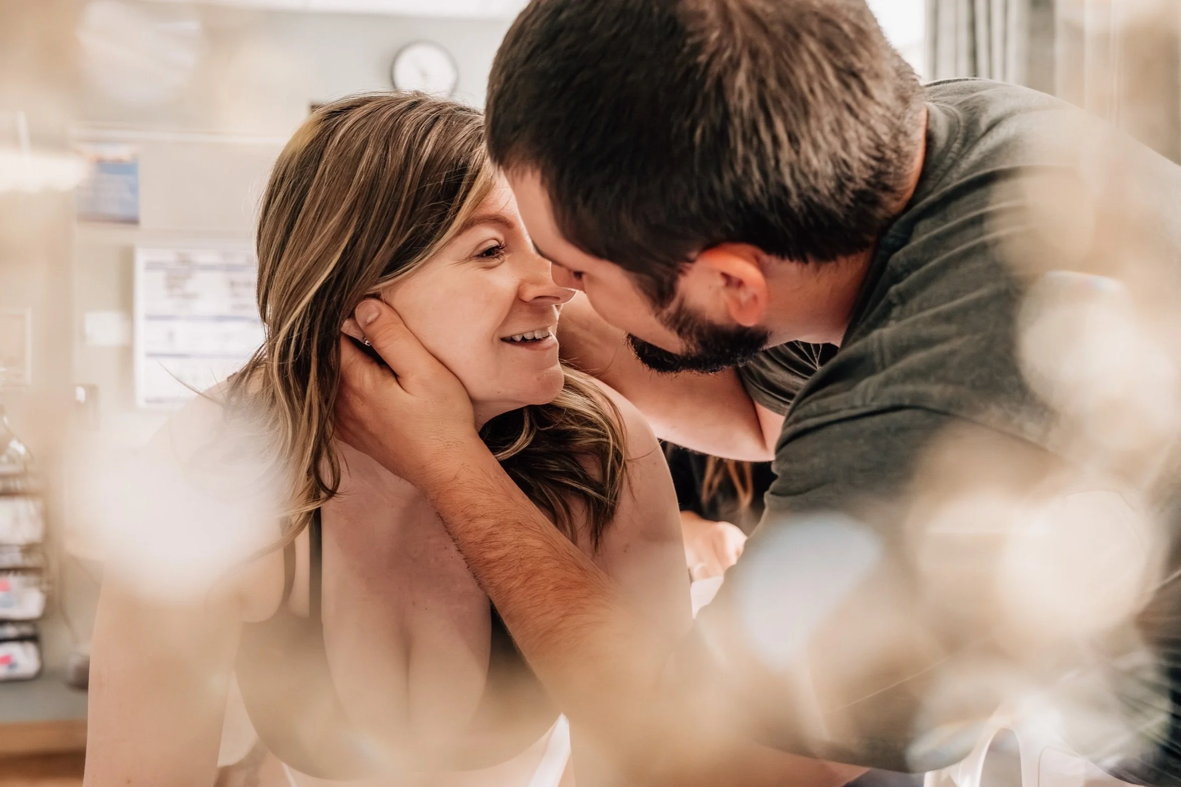 A husband leans in with his hands on his wife's face to kiss her. The woman, about to begin the pushing phase of labor, has a smile on her face and is surrounded by ethereal bokeh in the foreground of the photo.