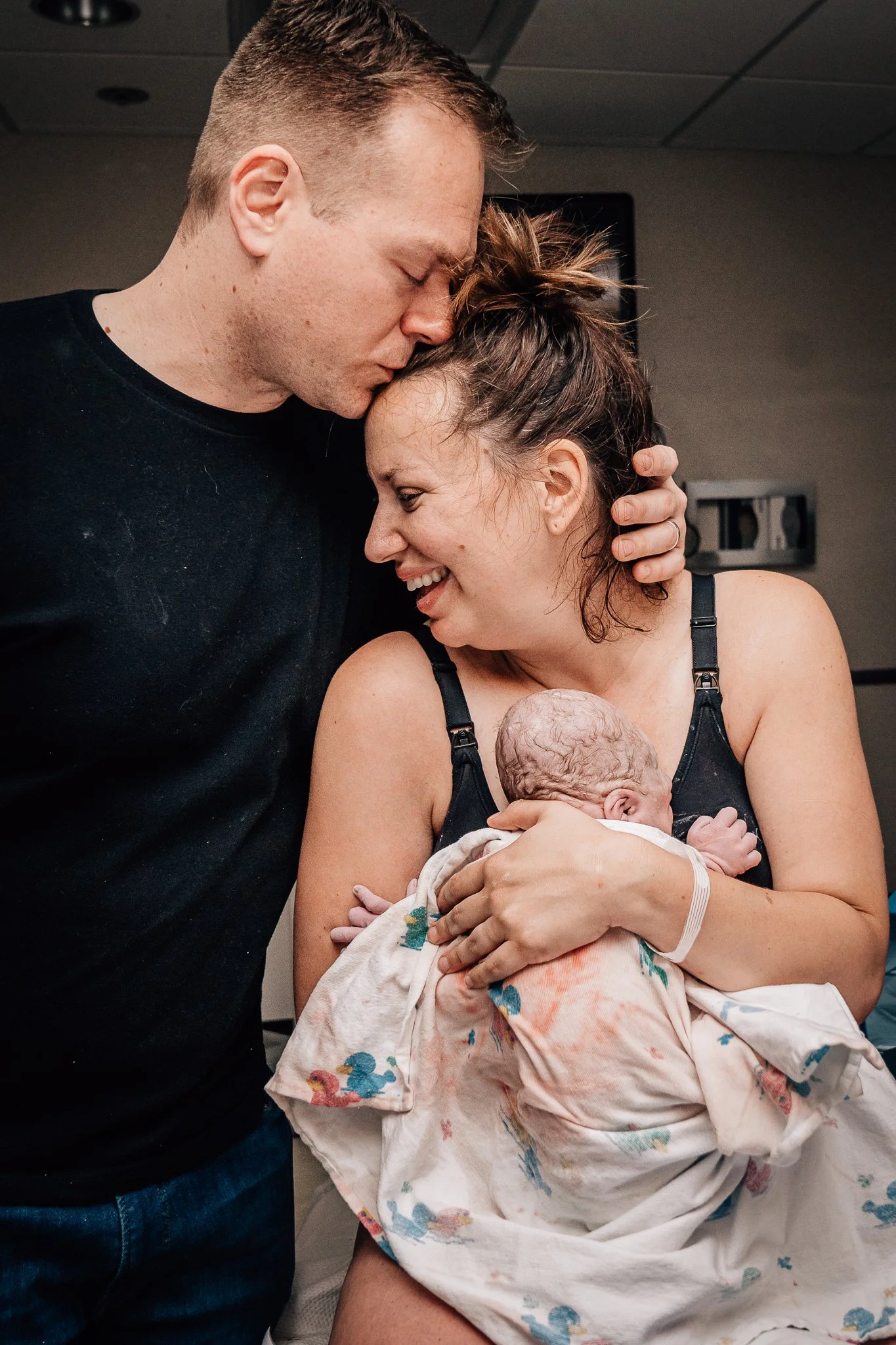 A smiling person holds their newborn baby to their chest moments after giving birth. Their partner has an arm around their shoulder and kisses their forehead in a moment filled with emotion. 