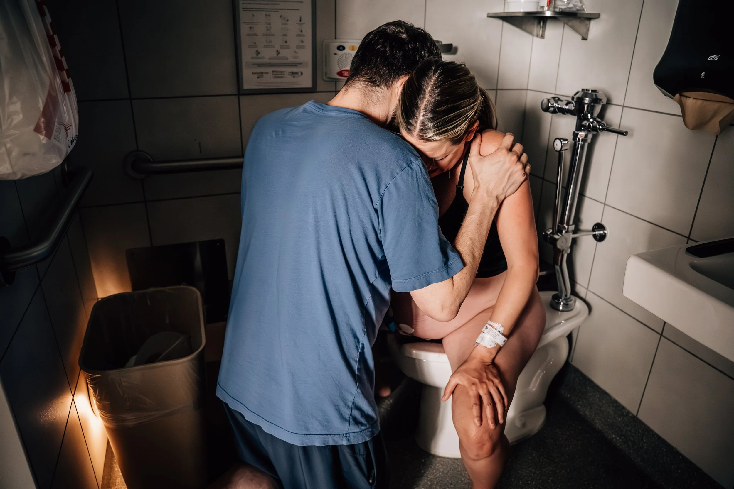 A woman in labor sits on the toilet and leans on her husband as he kneels in front of her, supporting her body and leaning into her with his hand on her shoulder. Light from outside streams in and creates dramatic lighting and shadows.
