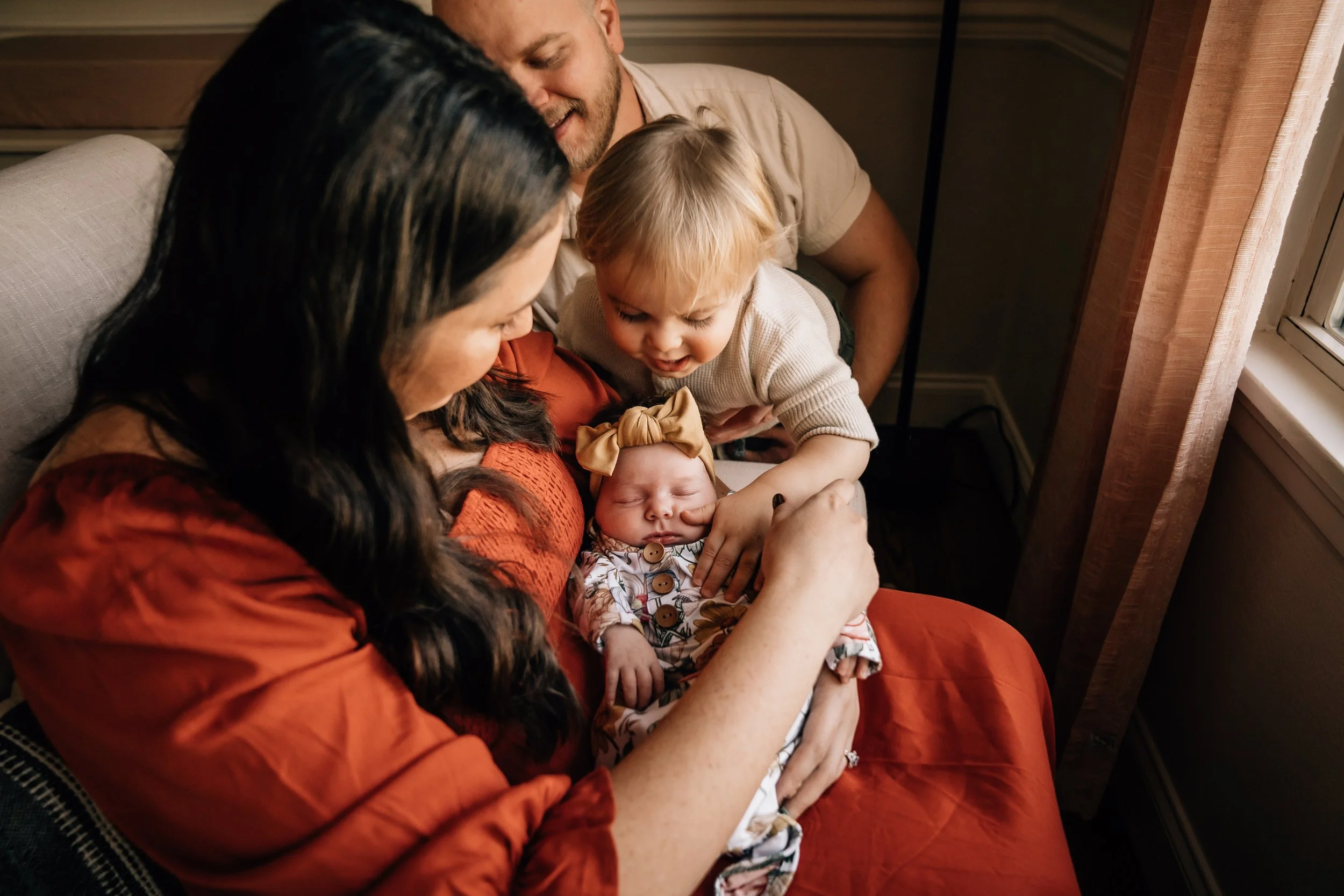 A mom wearing an orange dress sits in a rocking chair near a window cradling her newborn baby girl. The dad holds the toddler up to see the baby. 