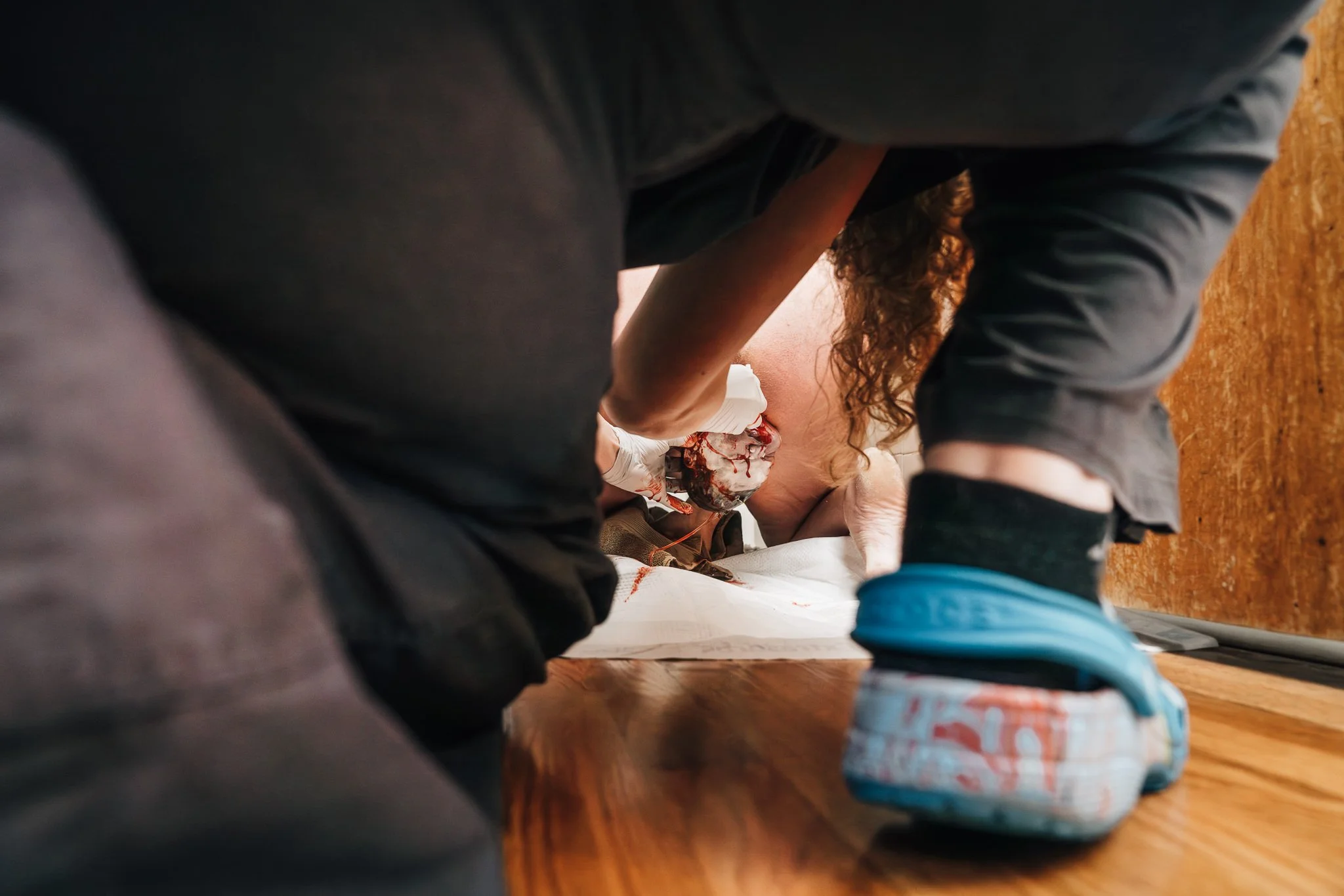 A midwife kneels on the floor, ready to help catch a baby being born at home. The baby’s head is emerging from the laboring parent.
