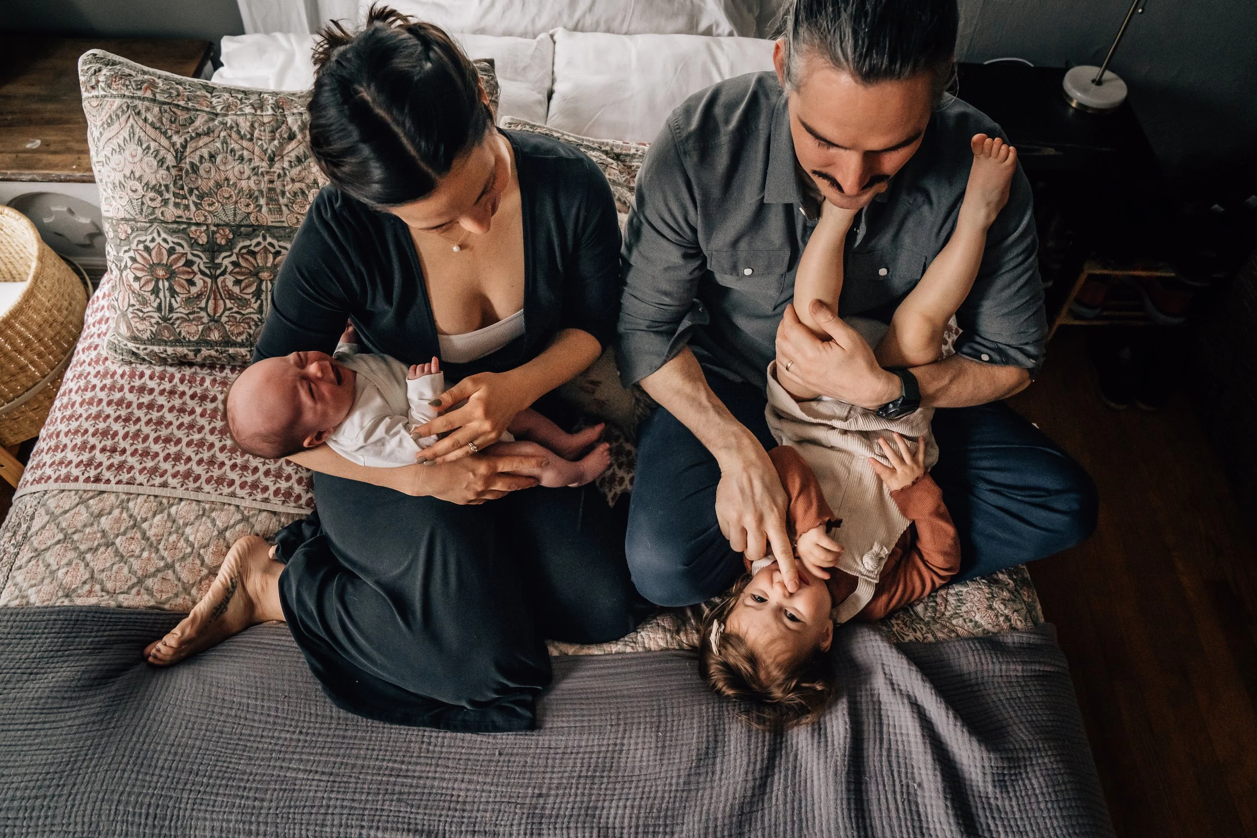 A family of four poses for a photo on their bed. The mom cradles the newborn baby and the dad holds the giggling toddler upside down.