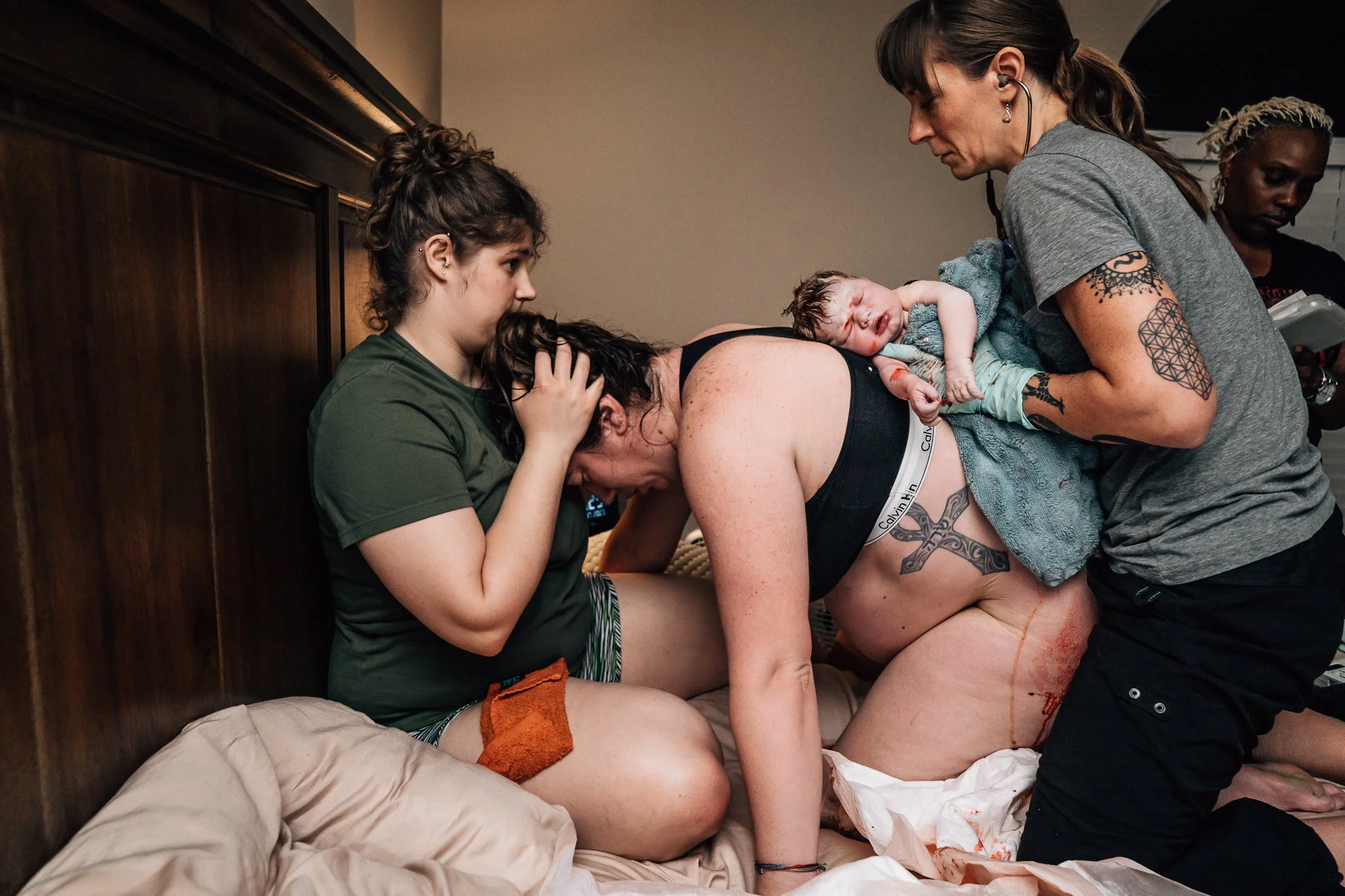 At a home birth, a midwife holds a newborn on the parent's back as the parent, who just gave birth in a hands-and-knees position on the bed, leans against their partner to recover.