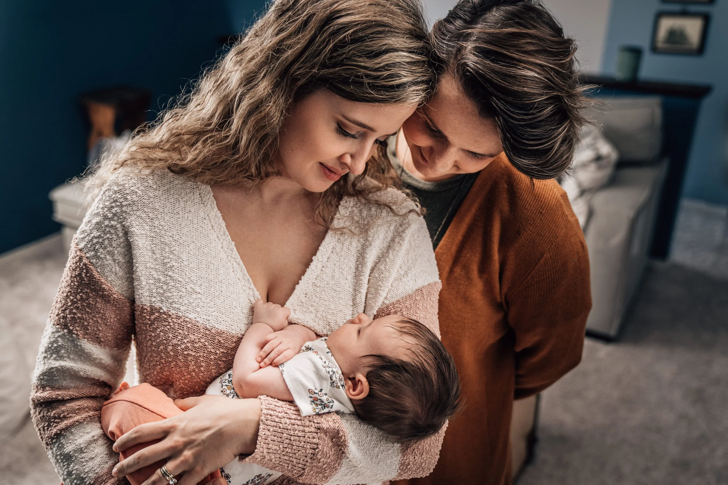 A couple wearing a cream sweater and an orange sweater stands together in their bedroom cradling their newborn baby girl.
