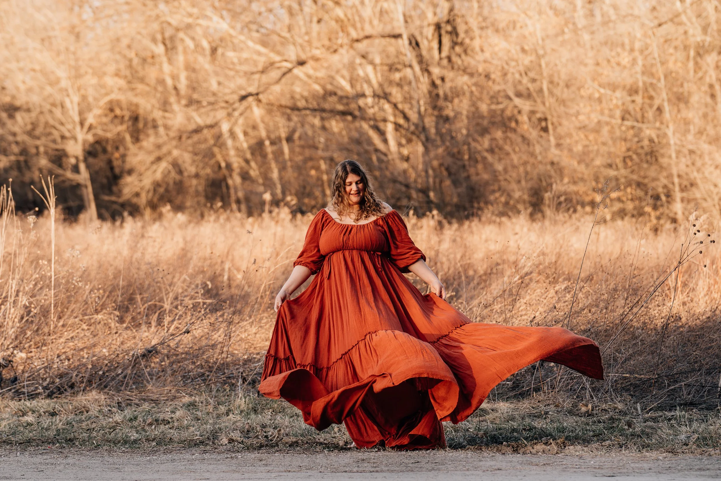 A pregnant woman in a flowing rust-colored dress stands on a dirt path, gracefully lifting the hem of her gown as it swirls around her. She looks down with a peaceful smile, surrounded by golden grasses and bare trees in soft evening light, creating