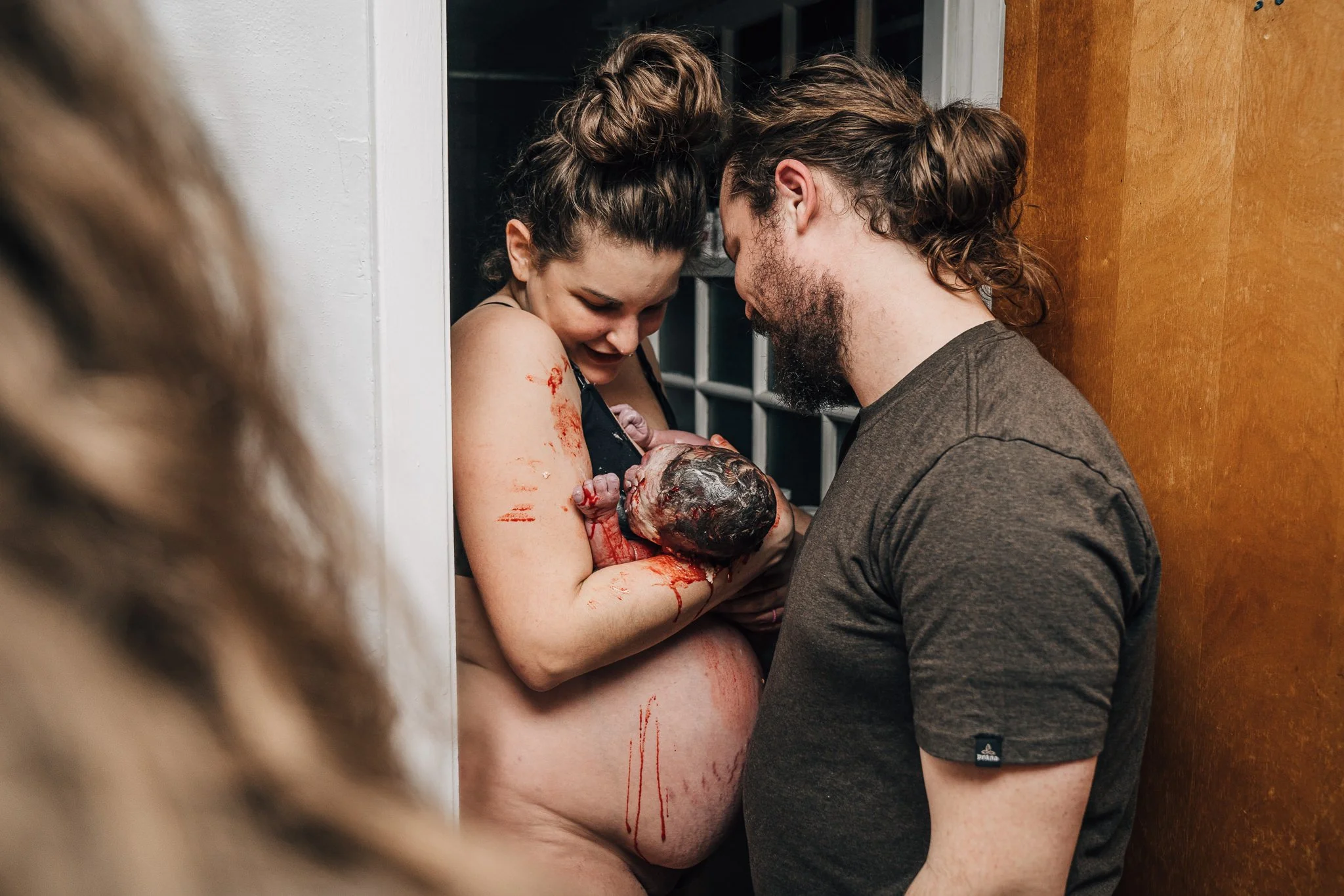 A person stands in the doorway of their bathroom, their partner beside them, smiling and holding their newborn baby just moments after birth.