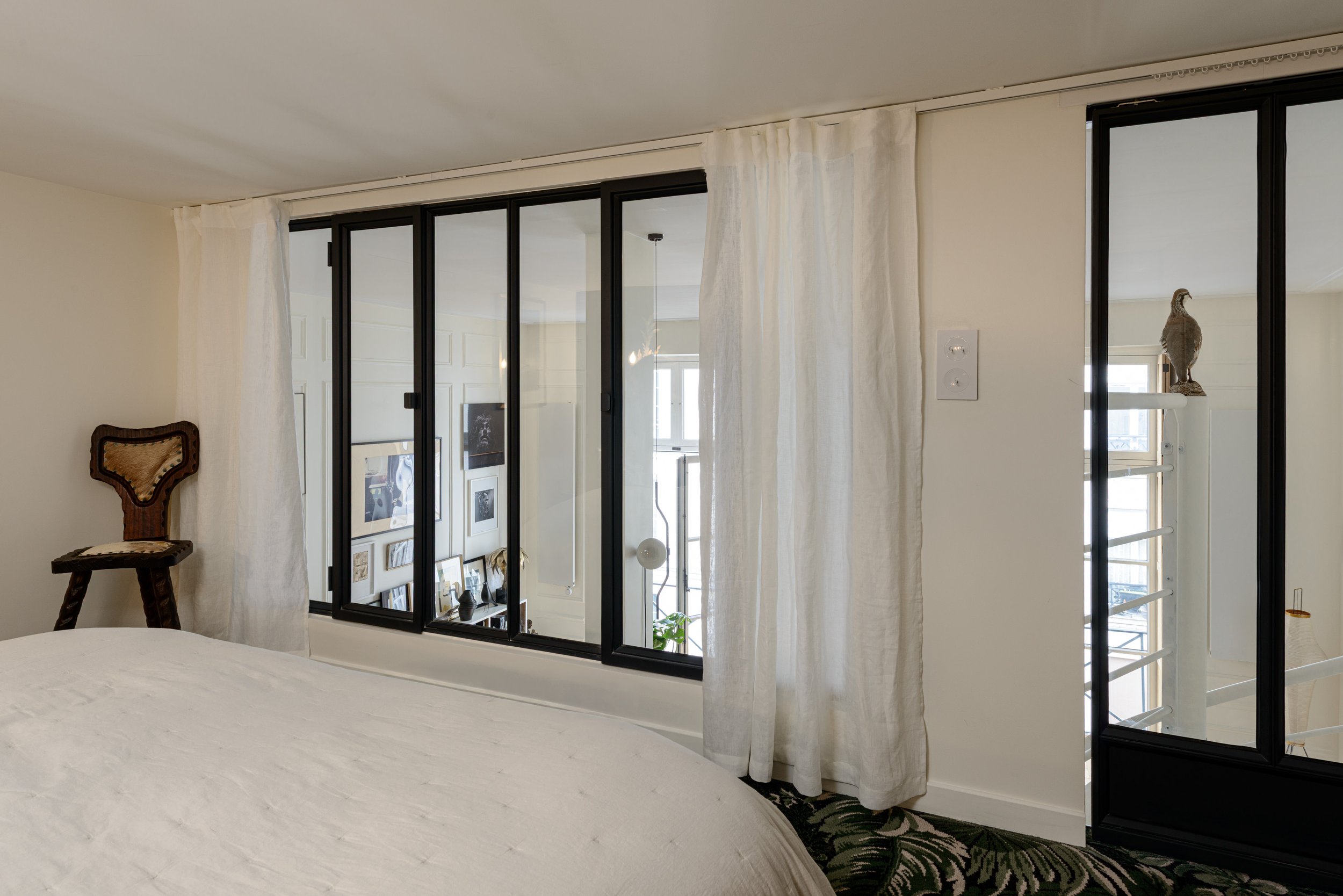 À l'étage, la chambre s’ouvre sur le séjour à travers une verrière. Au sol, la moquette fleurie apporte une touche vintage, comme un clin d’œil à la maison de Gainsbourg, rue de Verneuil.