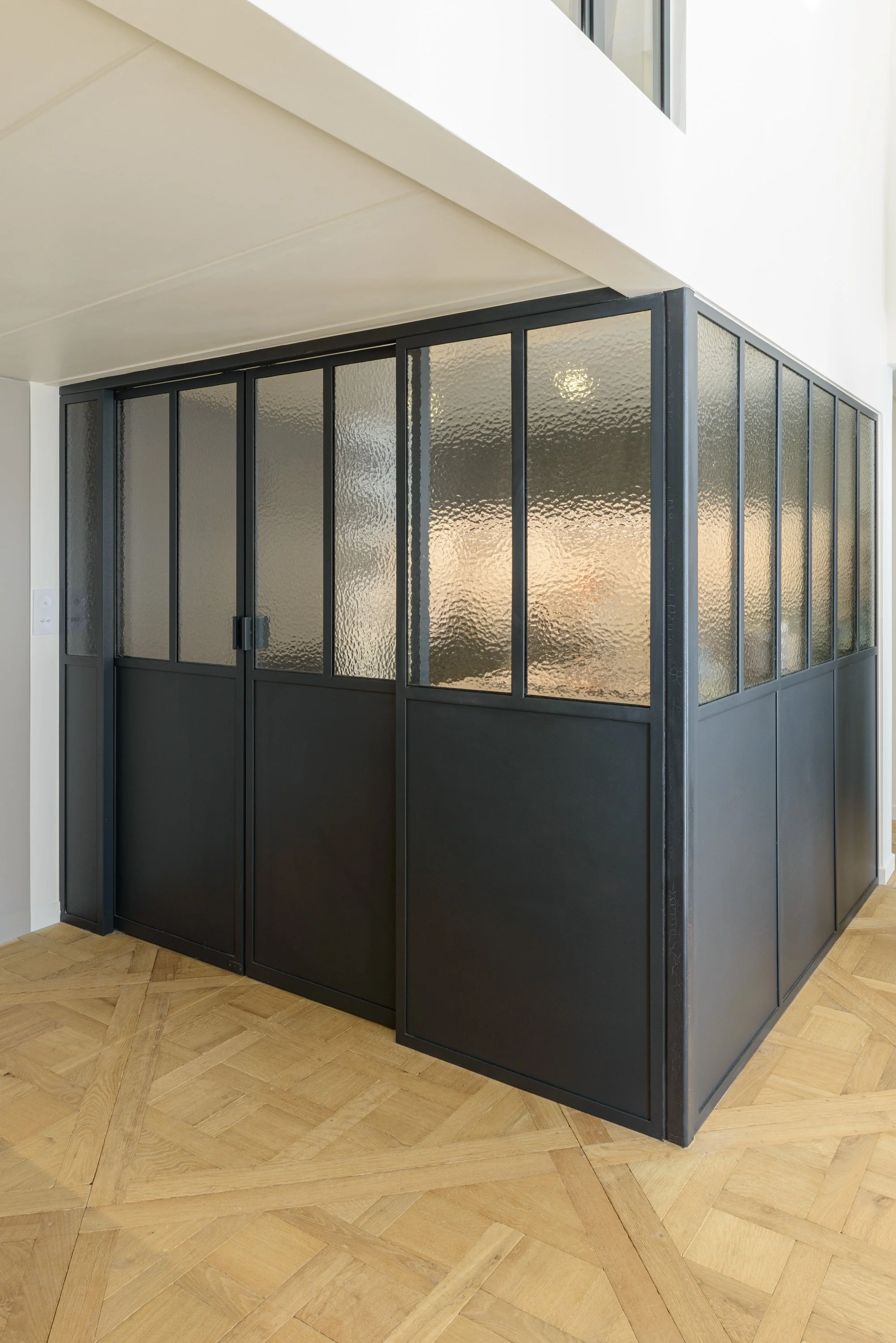 Fermée, la verrière isole la cuisine du séjour tout en laissant passer la lumière. Son dessin intègre discrètement la structure de la mezzanine, jusqu’au poteau dans l’angle.