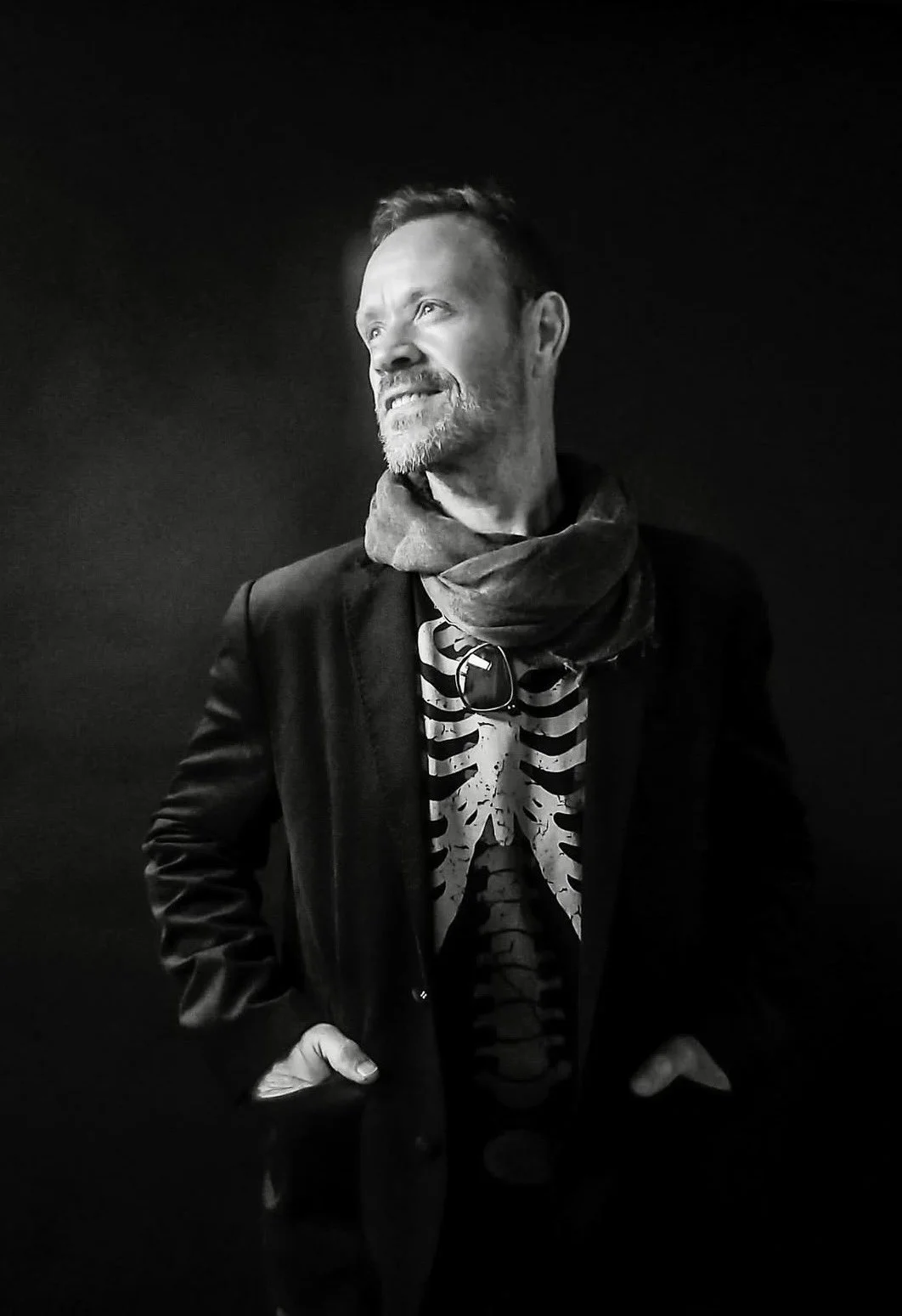Black and white photo of a man with short hair and beard, wearing a jacket, striped t-shirt with a chest skeleton outline design, and a scarf, standing against a dark background, looking to the left with a slight smile.