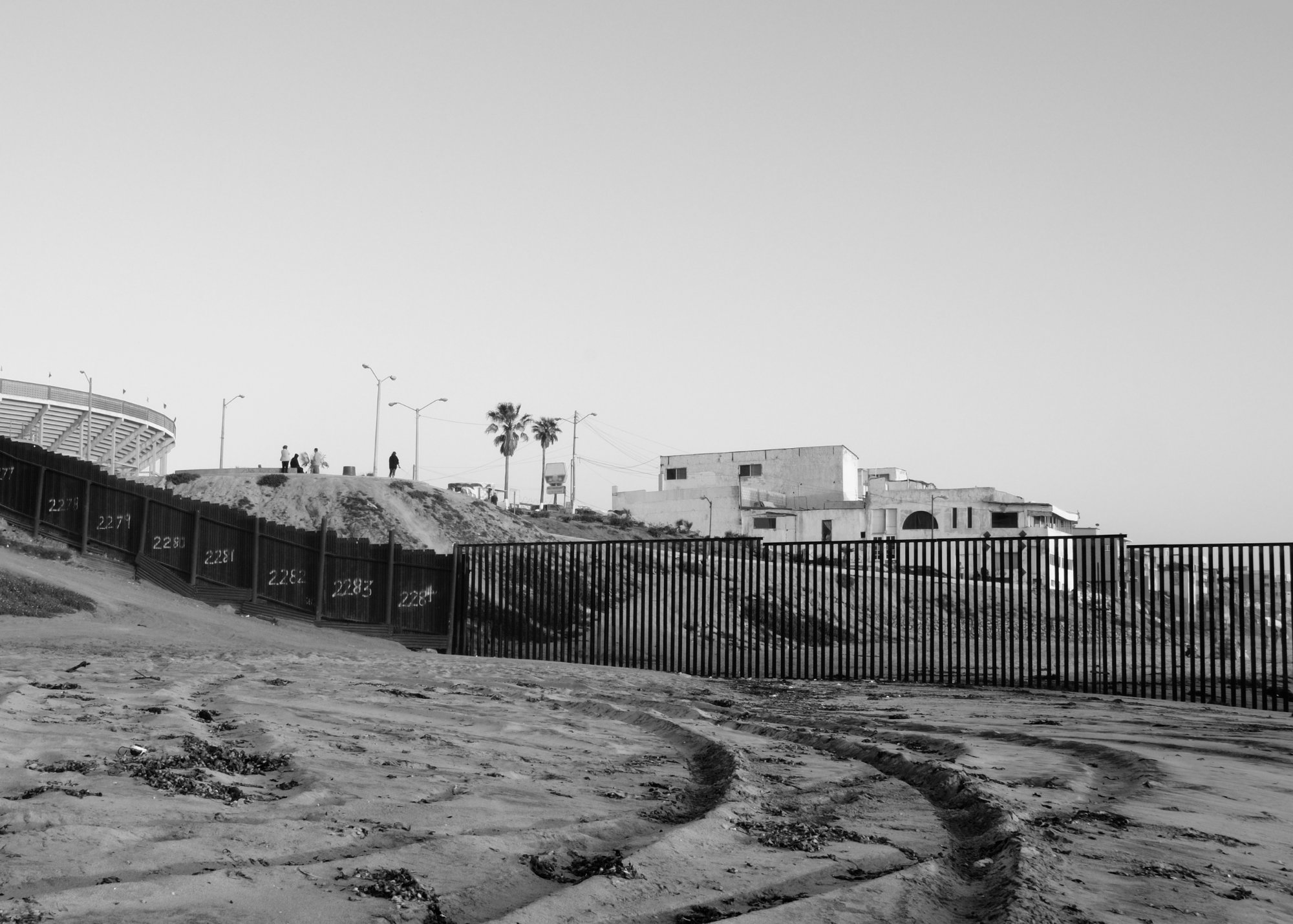 A country's border fence through a sandy area with buildings and palm trees behind the fence