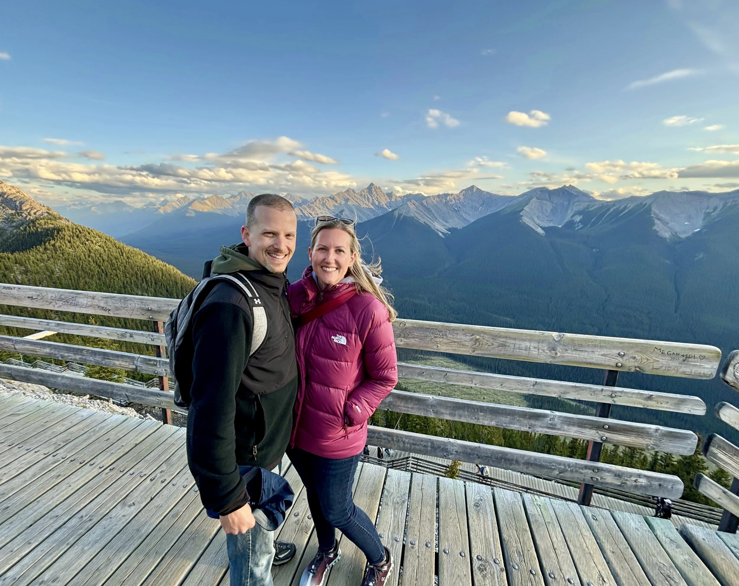 A smiling couple standing on a wooden observation deck with a mountain range in the background during sunset. The man is wearing a black jacket and has a backpack, the woman is wearing a purple jacket. Both are dressed for hiking.