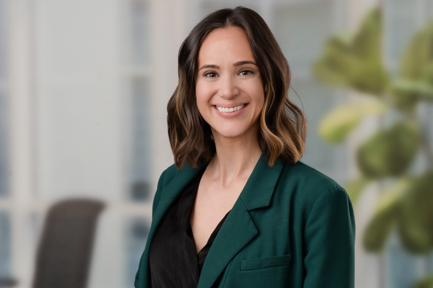 A woman with shoulder-length wavy brown hair, smiling, wearing a green blazer and black top, standing in an office environment.