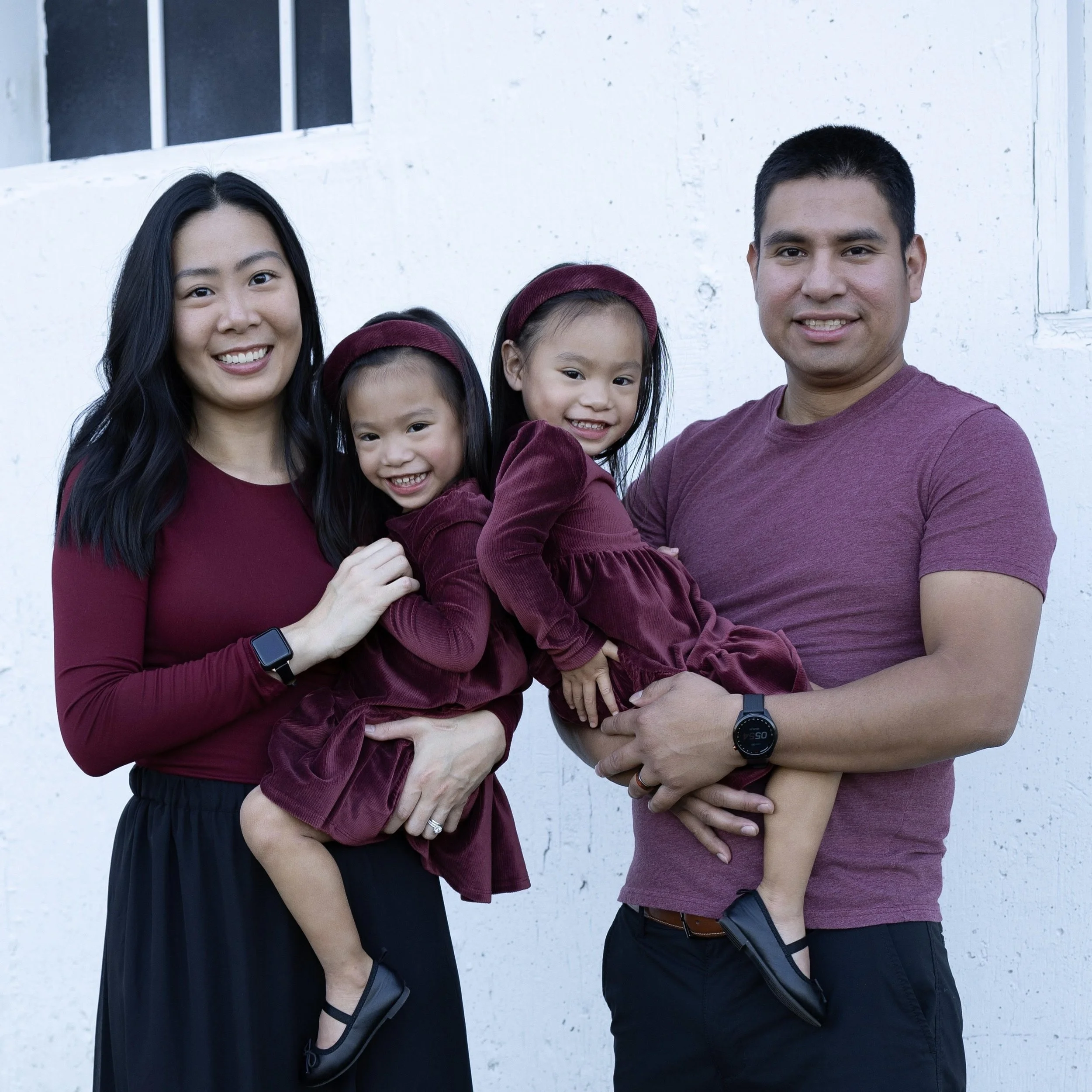 A family of four smiling and posing outdoors against a white wall. The mother and father each hold a young girl dressed in maroon. The mother has long black hair and wears a maroon top with a black skirt, while the father has short dark hair and wears a maroon T-shirt and black shorts. Both girls have maroon dresses and headbands.