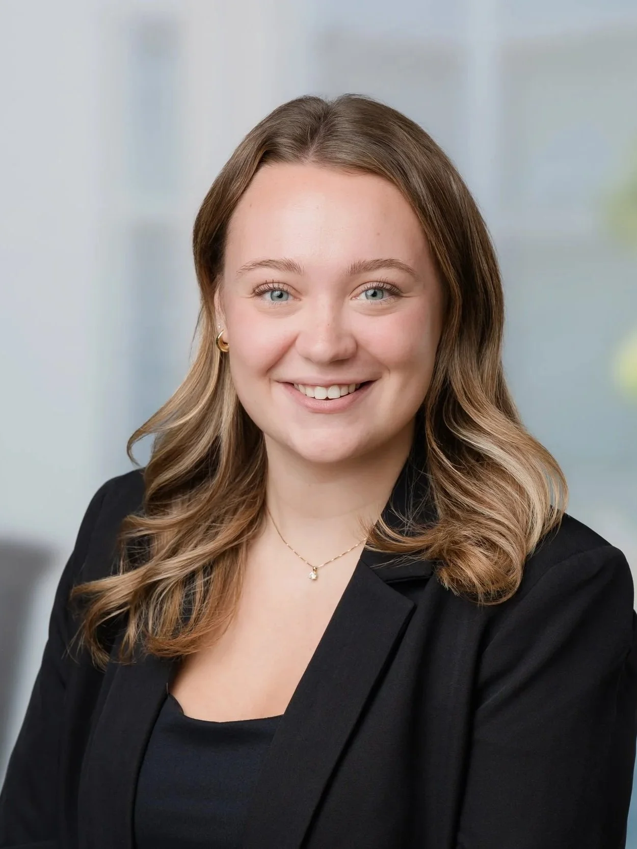 A smiling young woman with brown wavy hair, blue eyes, and gold jewelry, wearing a black blazer, in a professional setting with a blurred office background.