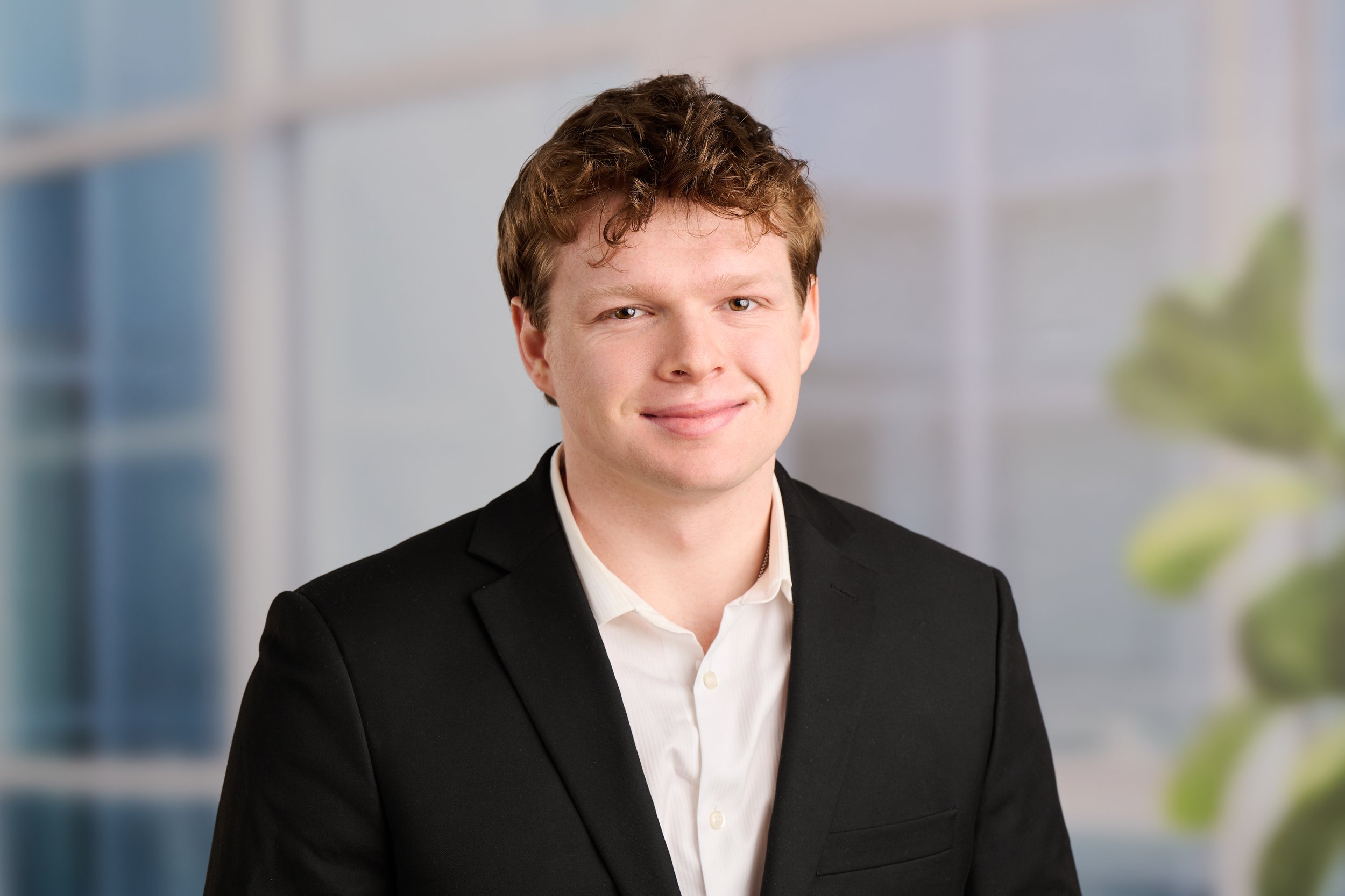 A young man with light brown, curly hair and fair skin wearing a black suit jacket and white shirt, standing indoors with a blurred office background.