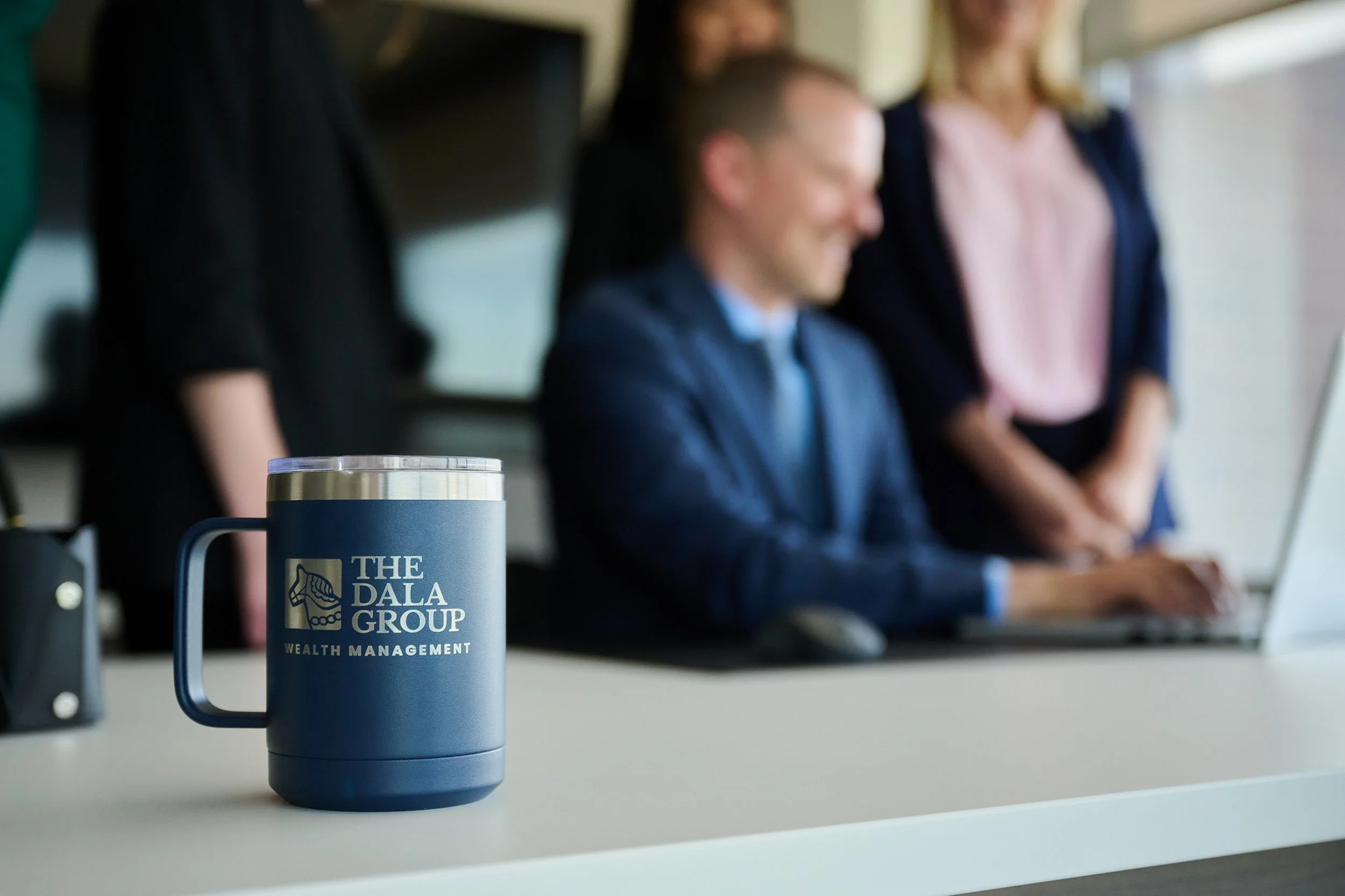 Close-up of a blue coffee mug with the logo of The Dala Group, a wealth management company, placed on a white table. In the background, blurred people are sitting and standing, engaged in a meeting or discussion in a modern office space.
