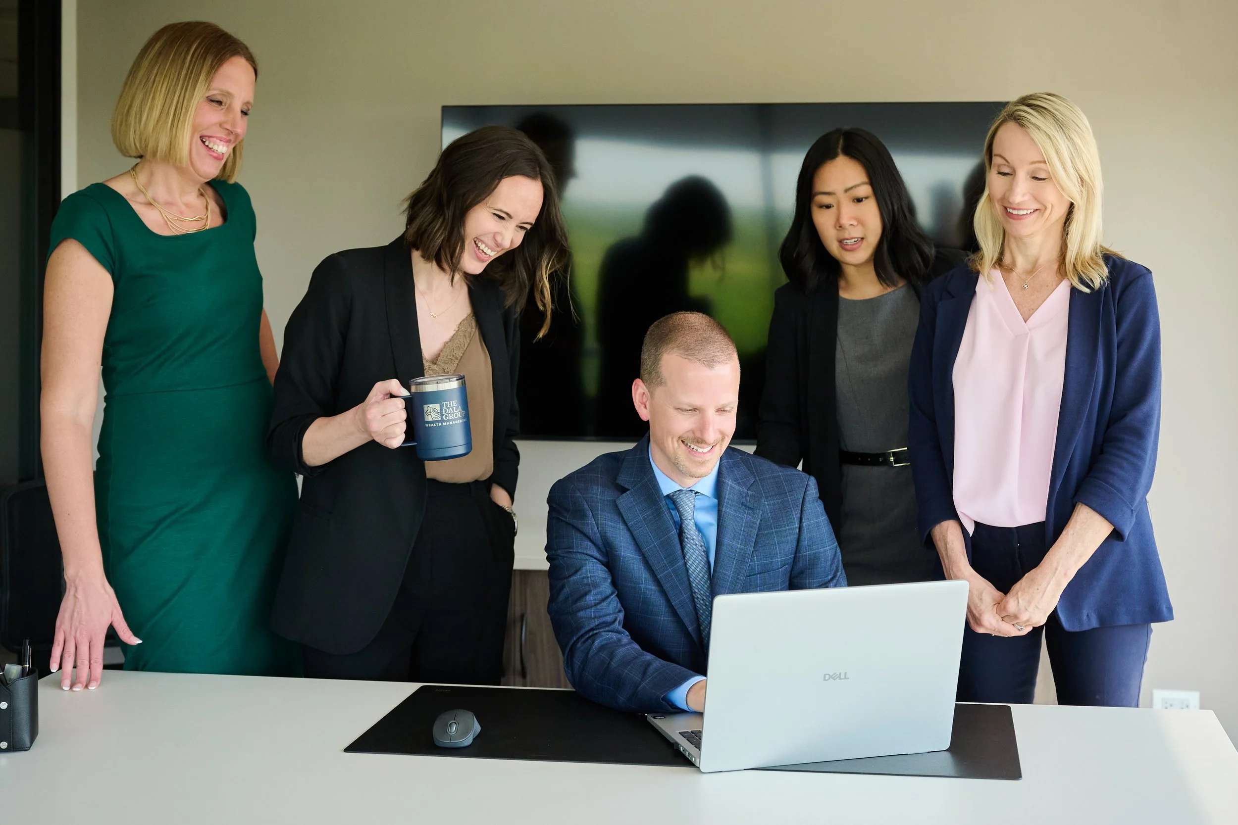 Group of five professional people gathered around a man working on a laptop in an office setting, with some smiling and others watching.